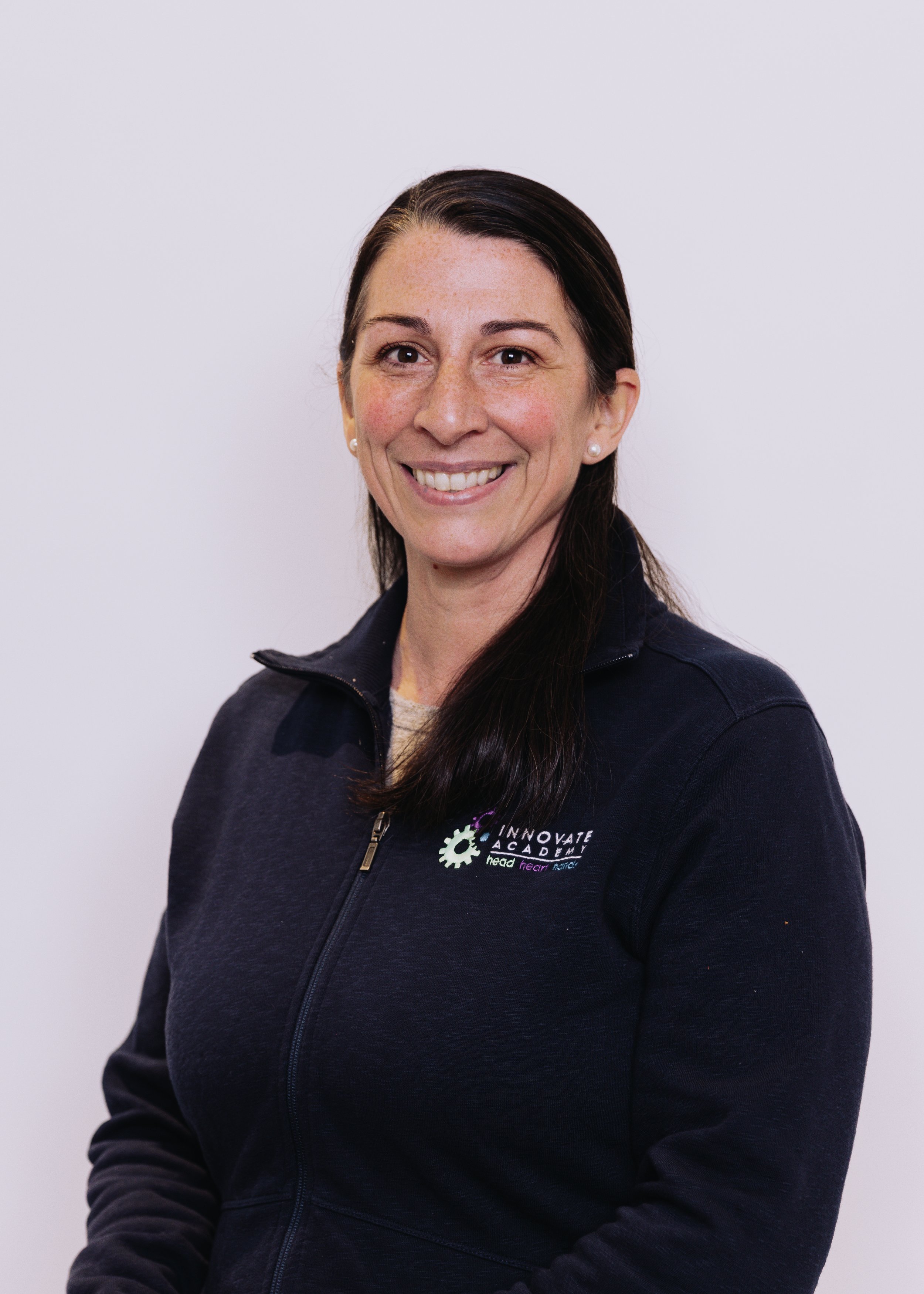 A woman with long dark hair, wearing a dark navy zip-up jacket with a logo on the chest that says 'Innovate Academy'. She is smiling and standing against a plain light background.