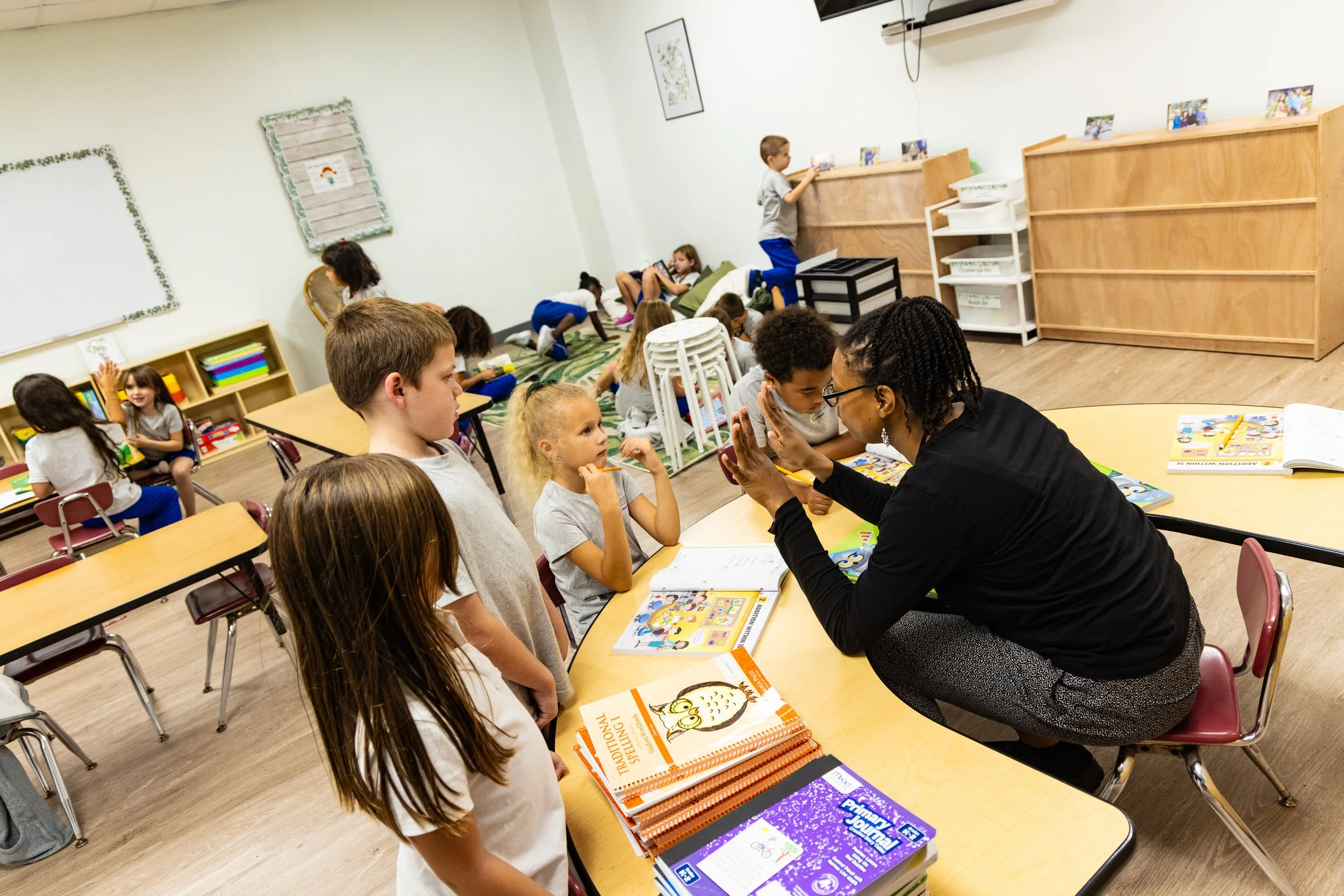 A teacher sitting at a table with young students in a classroom, showing a hand gesture to a girl standing beside her, with other children engaged in activities in the background.
