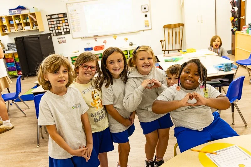 Six young children, five girls and one boy, are standing together in a classroom, smiling and making heart shapes with their hands. Other children are seated at tables, with classroom supplies and educational posters visible in the background.