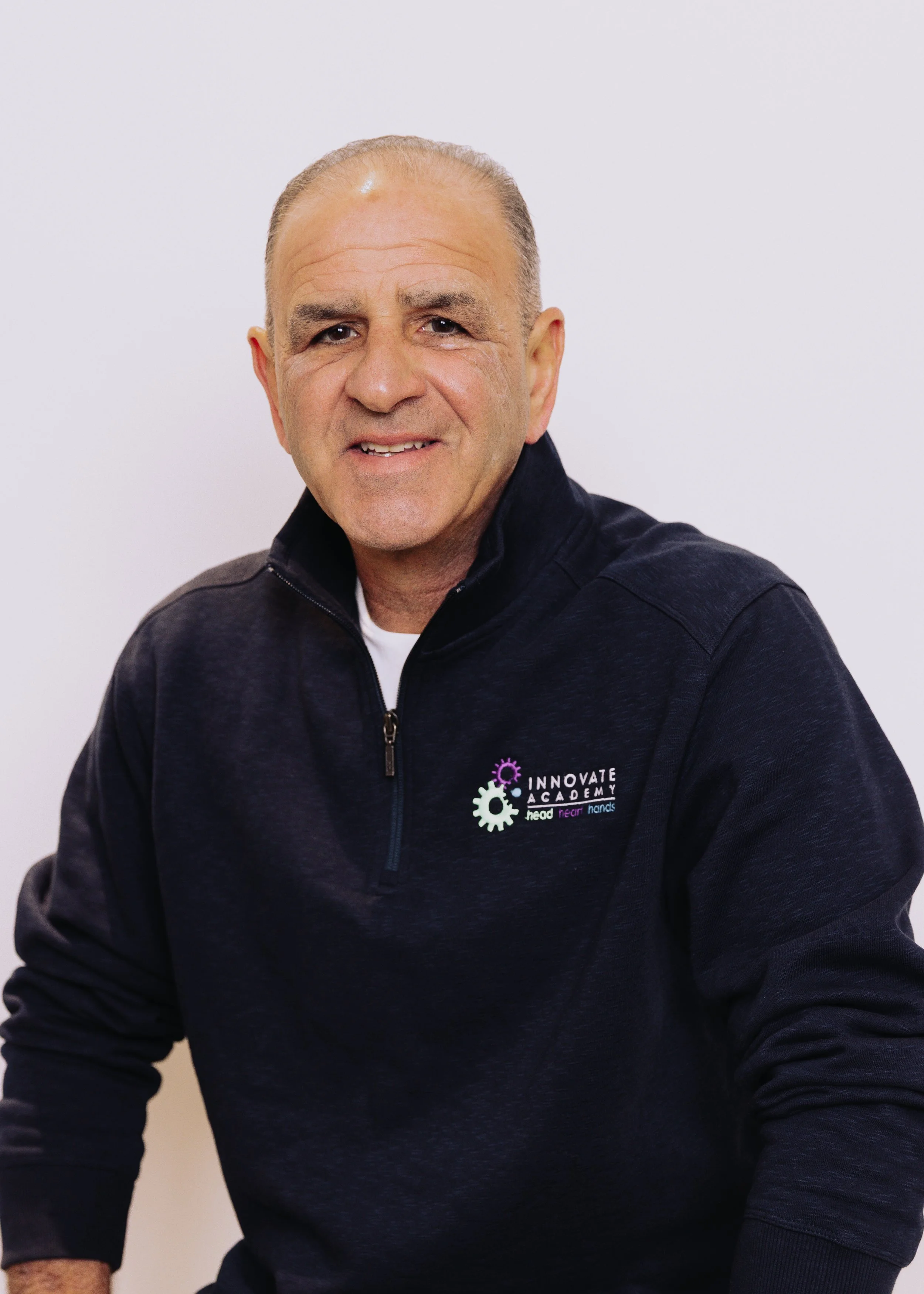A middle-aged man with short gray hair, wearing a black zip-up jacket with a logo that reads "Innovate Academy" and a white shirt underneath, smiles at the camera against a plain white background.