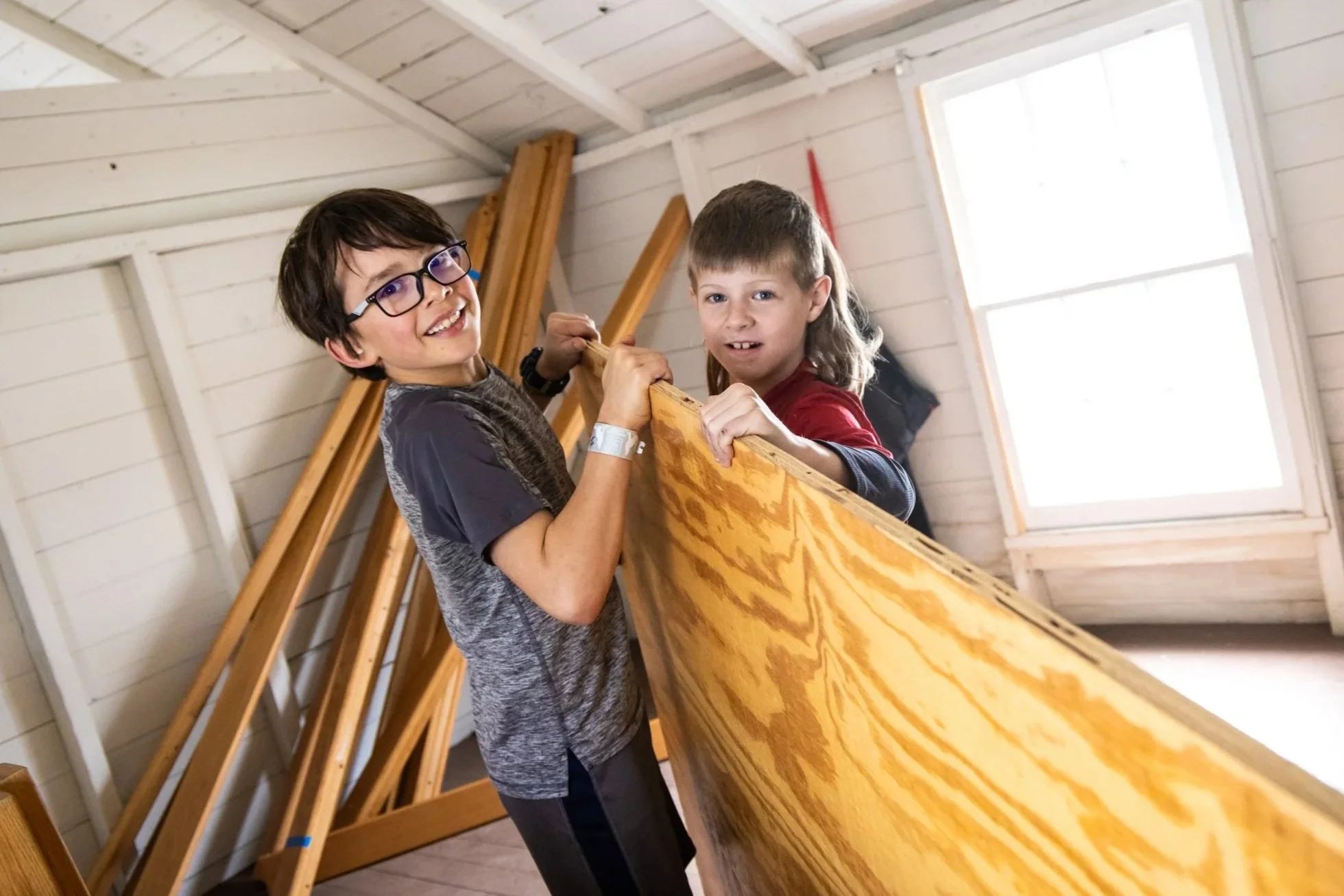 Two children holding a large piece of plywood inside a woodworking shop, with wooden beams and windows in the background.