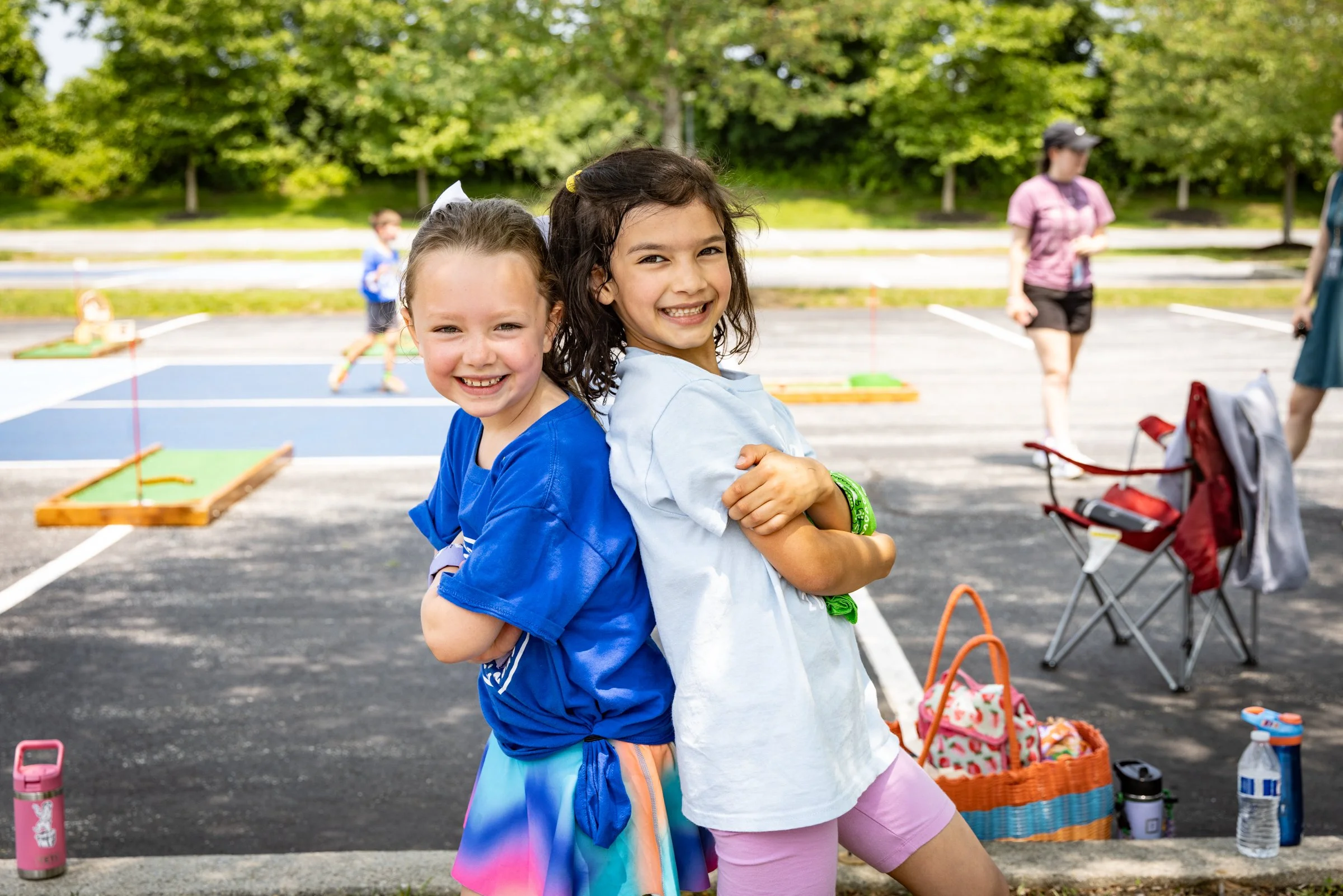 Two young girls smiling and posing back-to-back outdoors near a parking lot with shuffleboard courts, surrounded by trees and other children and adults in the background.