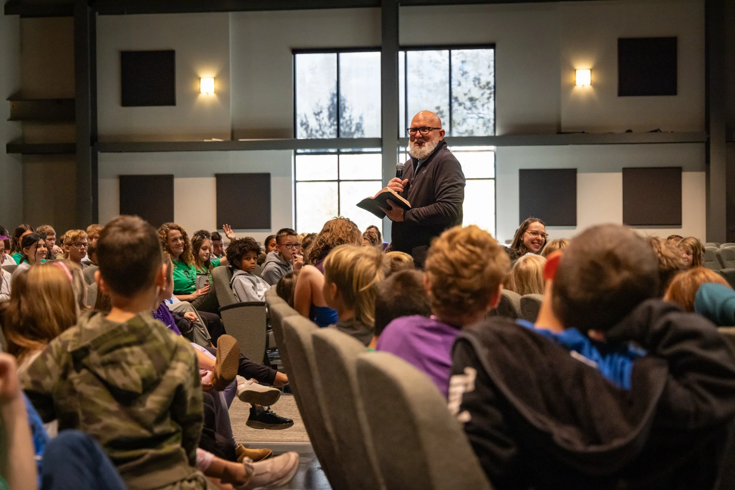 A man with glasses and a beard stands on stage holding a microphone and a book, addressing a seated audience of children and teenagers in a large room with high windows and acoustic panels.