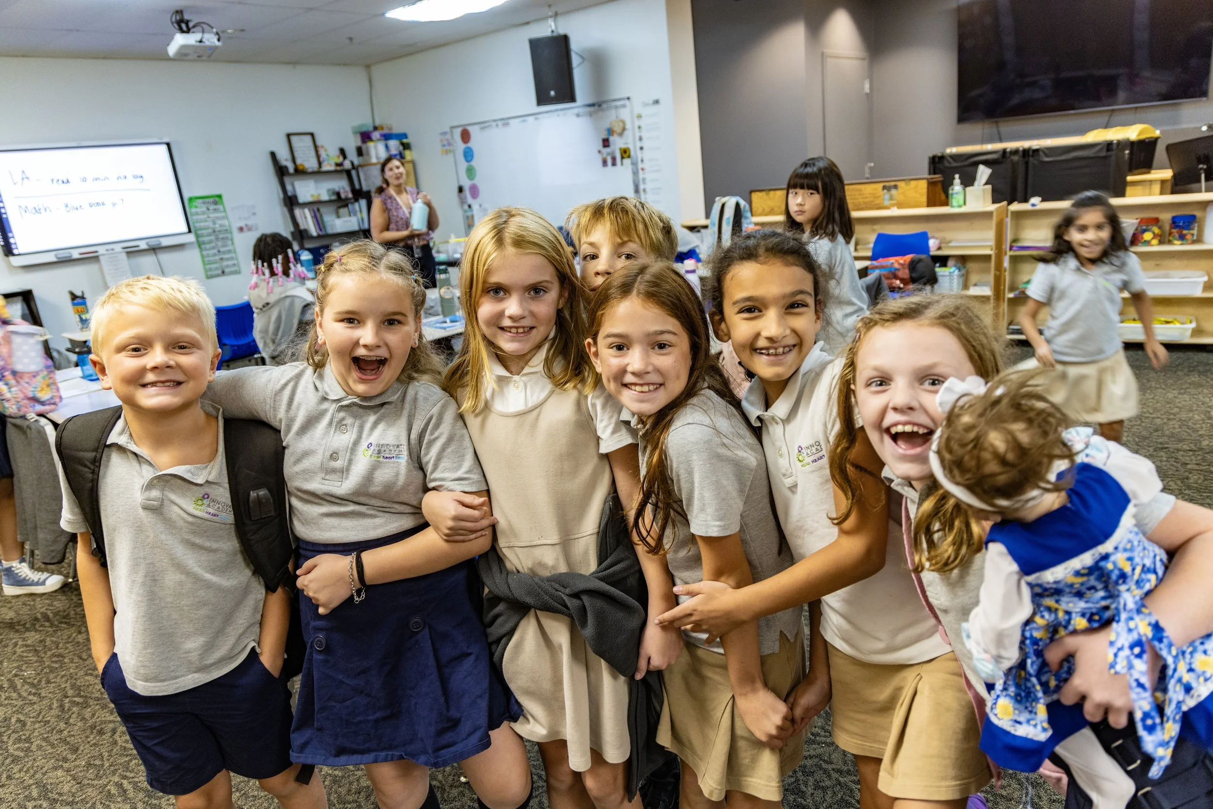 Group of smiling young children in school uniform in classroom, some with backpacks, standing together for photo, with whiteboard and teacher in background.