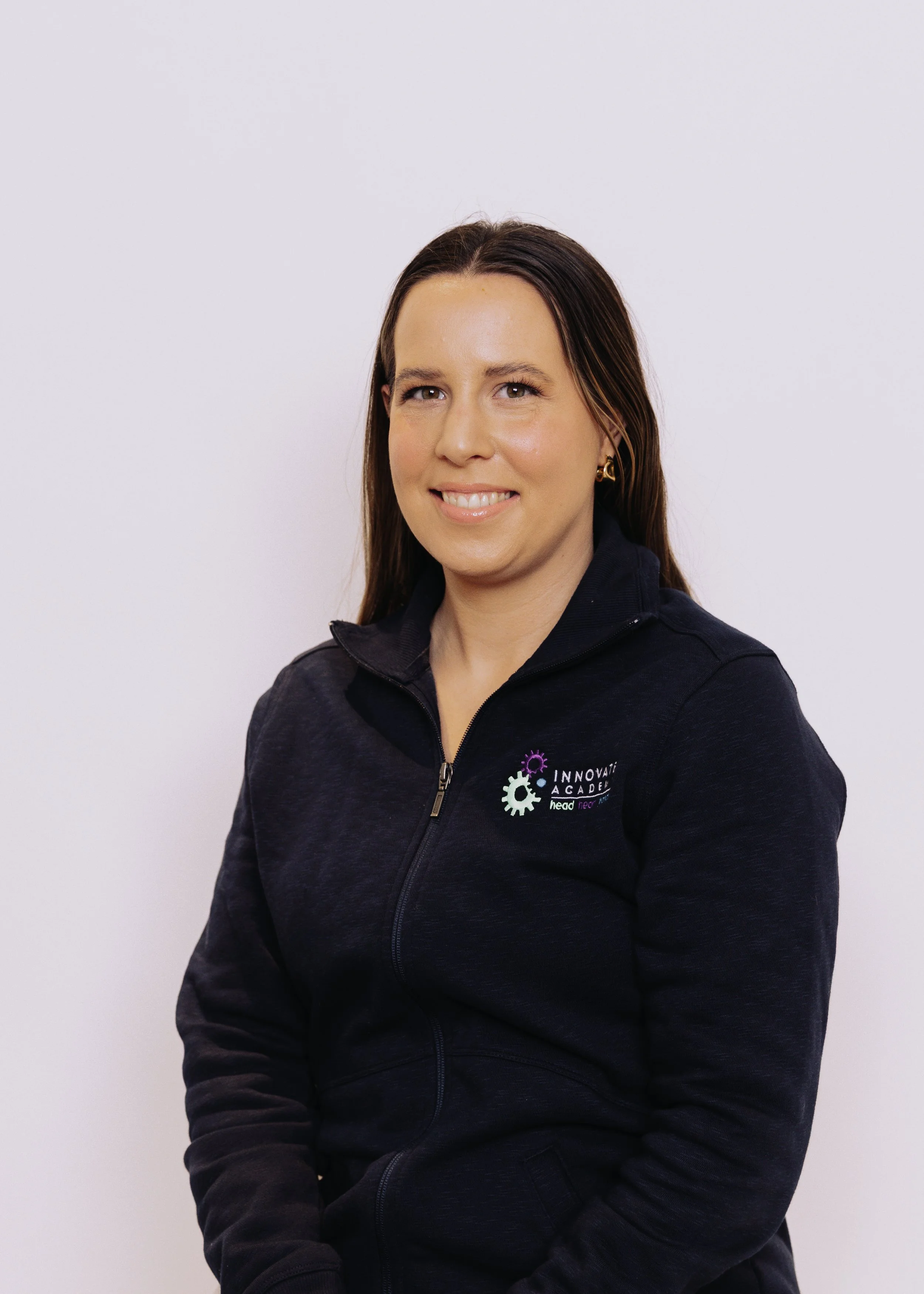 A woman with brown hair smiling, wearing a navy blue jacket with a logo on the chest that says Innovation Academy.