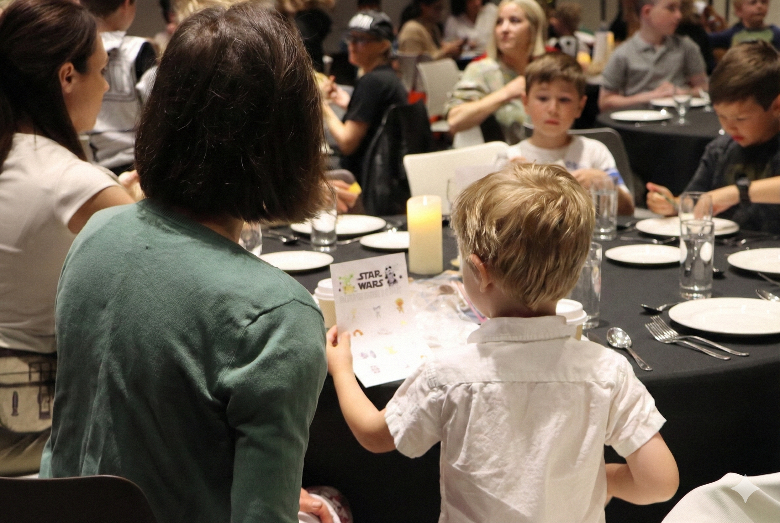 A woman and a young boy sitting at a round dining table with a Star Wars-themed placard, a candle, and dinnerware, attending a group event or dinner party.
