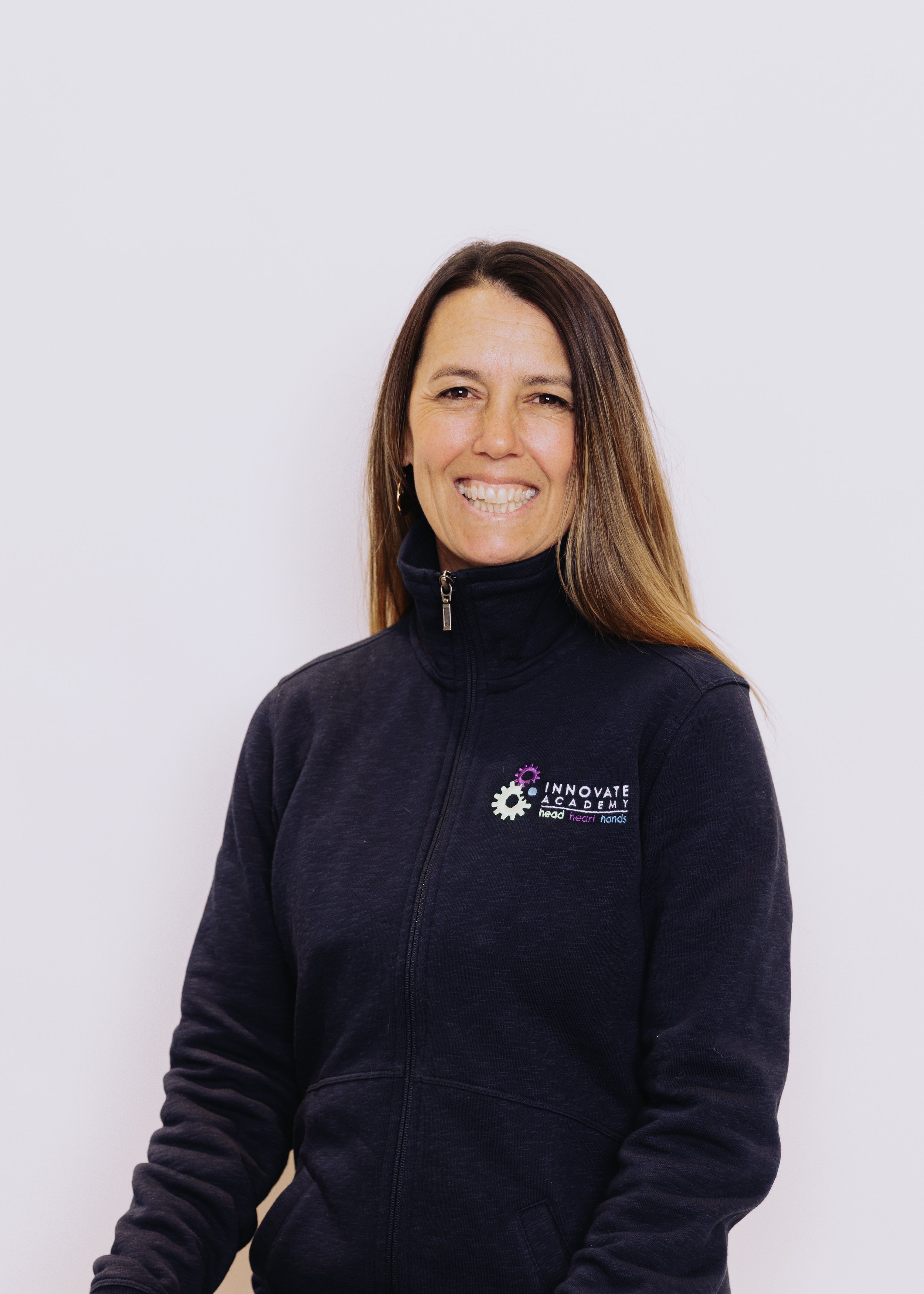 A woman with brown hair smiling, wearing a black zip-up jacket with the logo 'Innovate Academy' on the chest, standing against a plain white background.