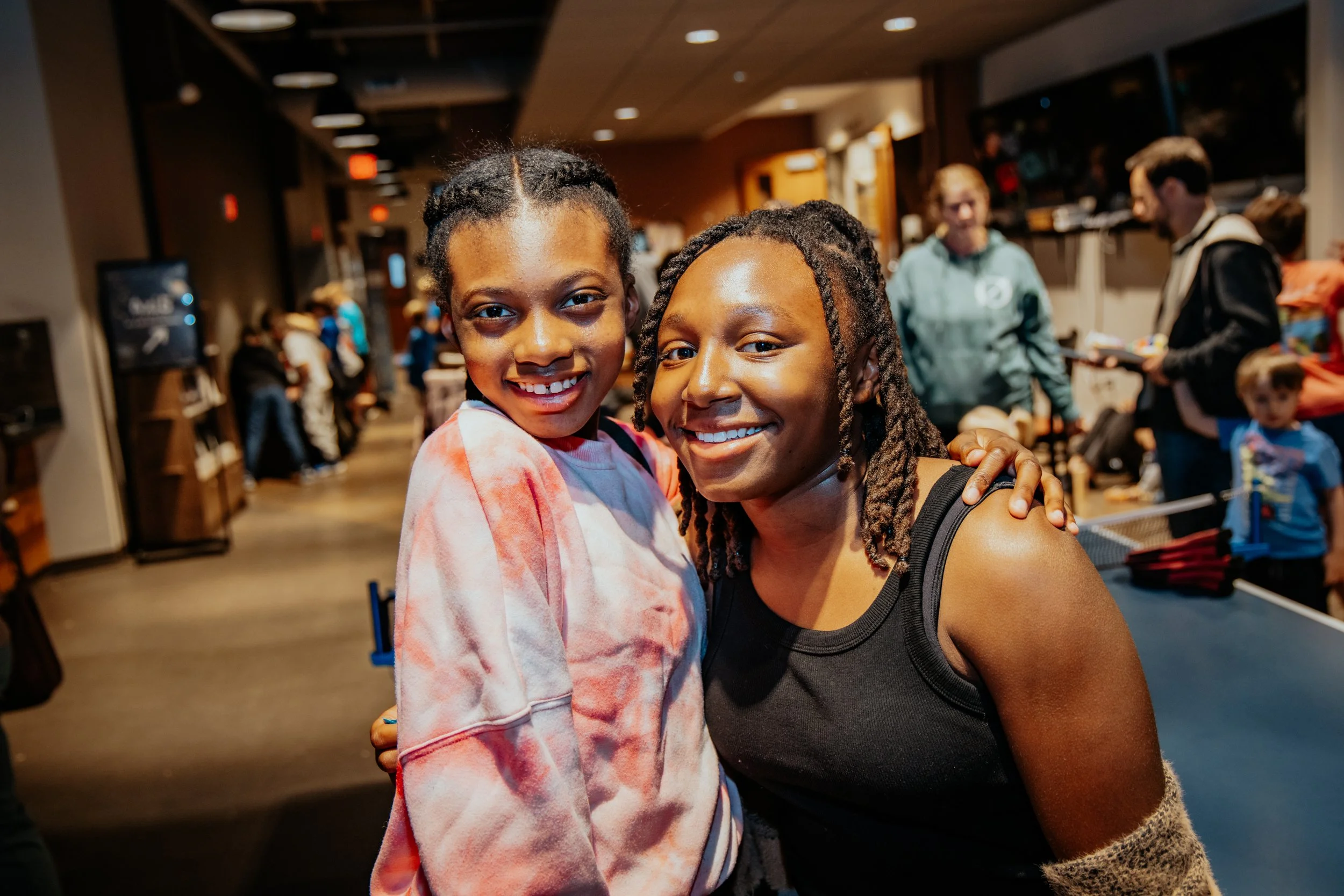 Two smiling women with dark hair and brown skin, one wearing a tie-dye hoodie and the other in a black tank top, standing close together in a busy indoor social setting with people in the background.