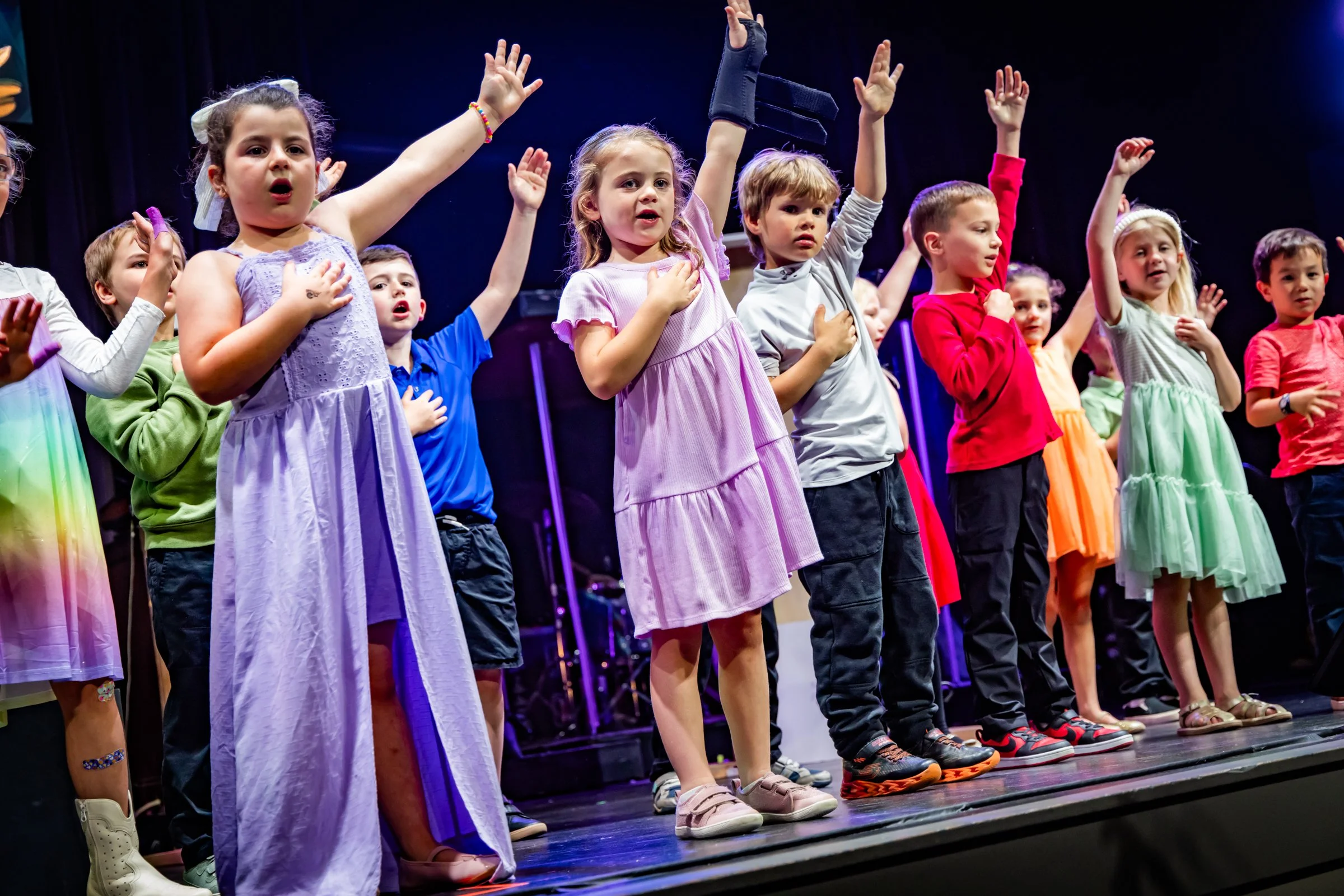 Children singing on stage with their right hands over their hearts during a performance in colorful clothing.