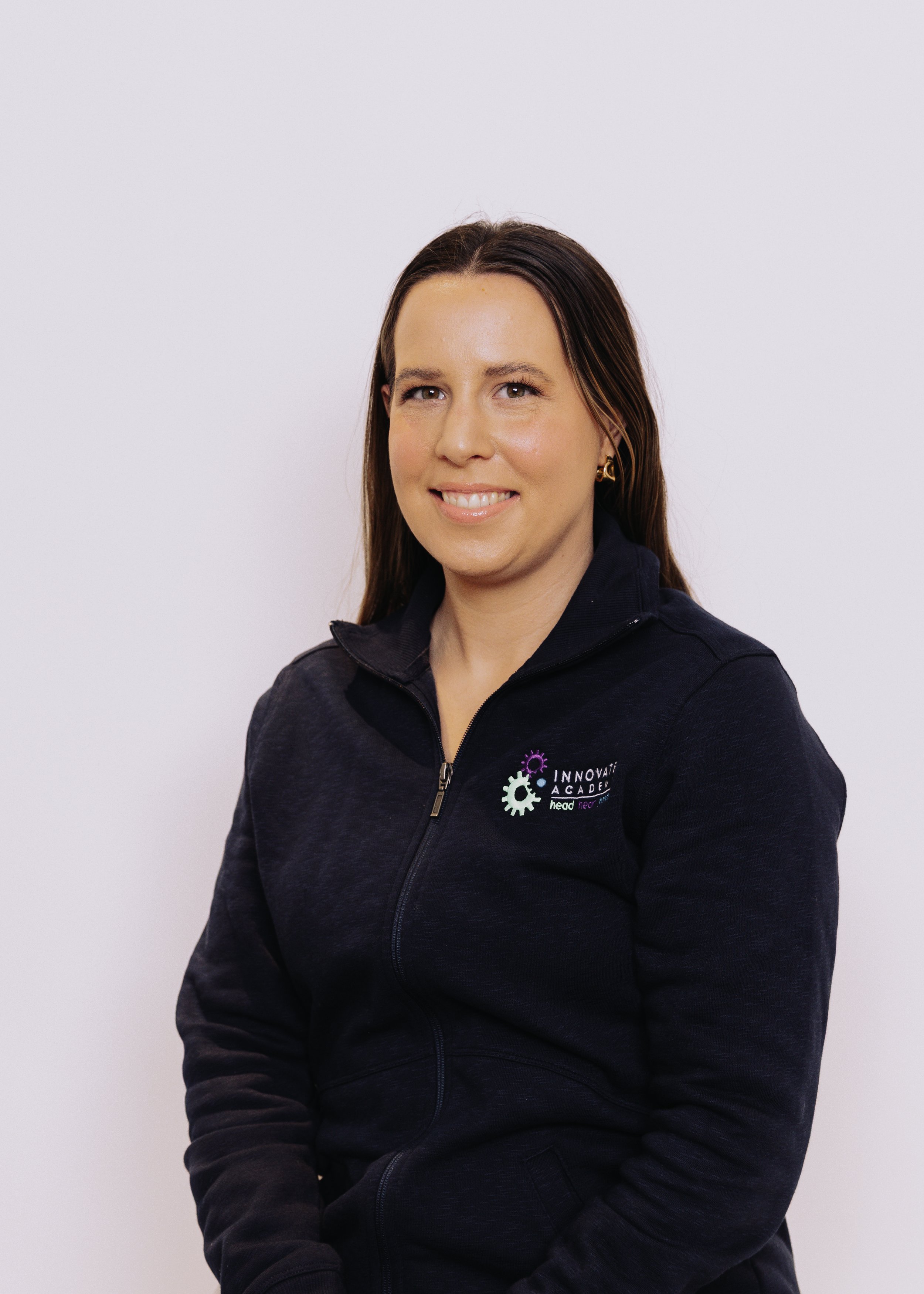 A woman with long brown hair smiling, wearing a black zip-up jacket with a logo that says 'Innovative Academy' on the chest, against a plain white background.