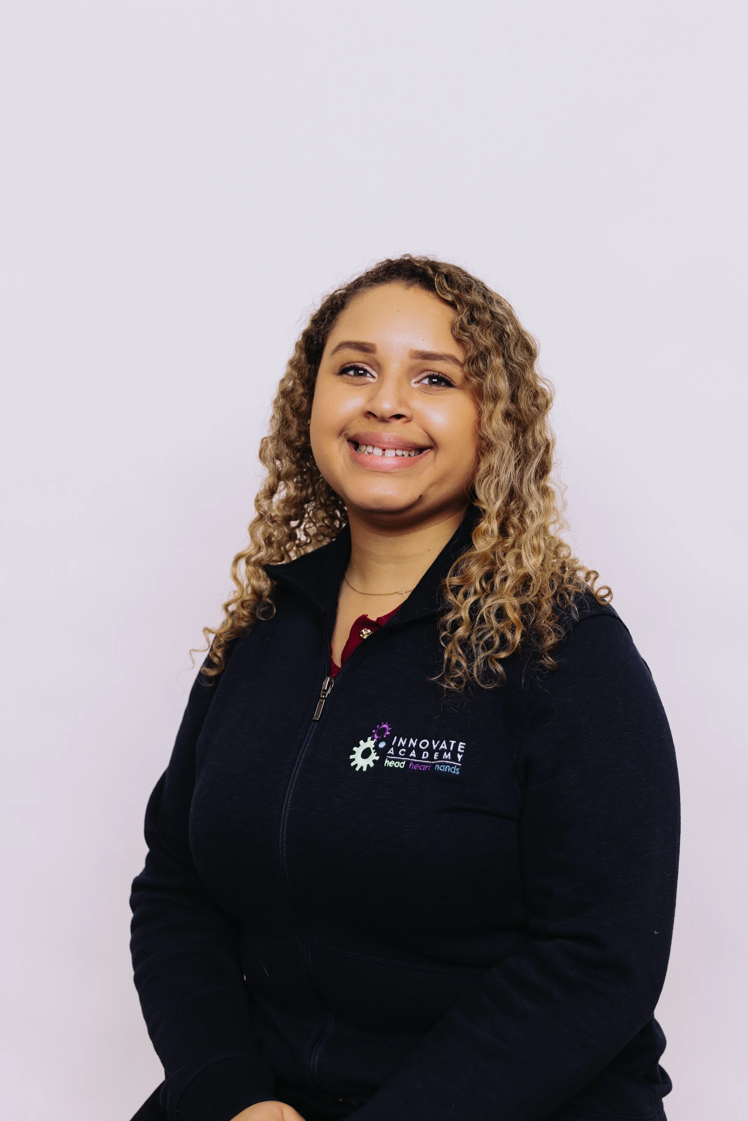 A young woman with curly blonde hair, wearing a black top and a navy blue jacket with a logo, standing against a plain white background.