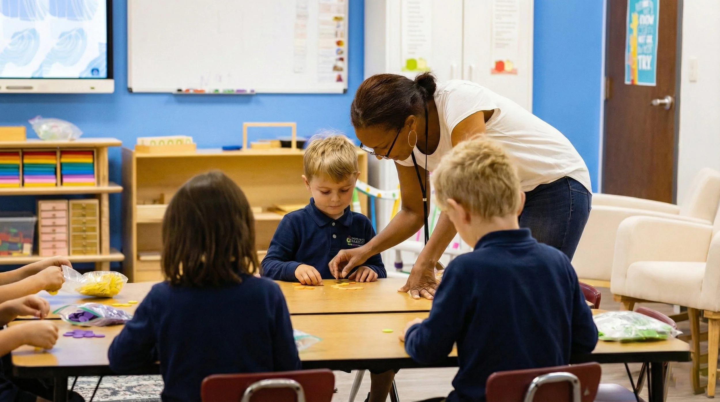An adult woman teaching children at a table in a classroom, with educational posters and shelves in the background.