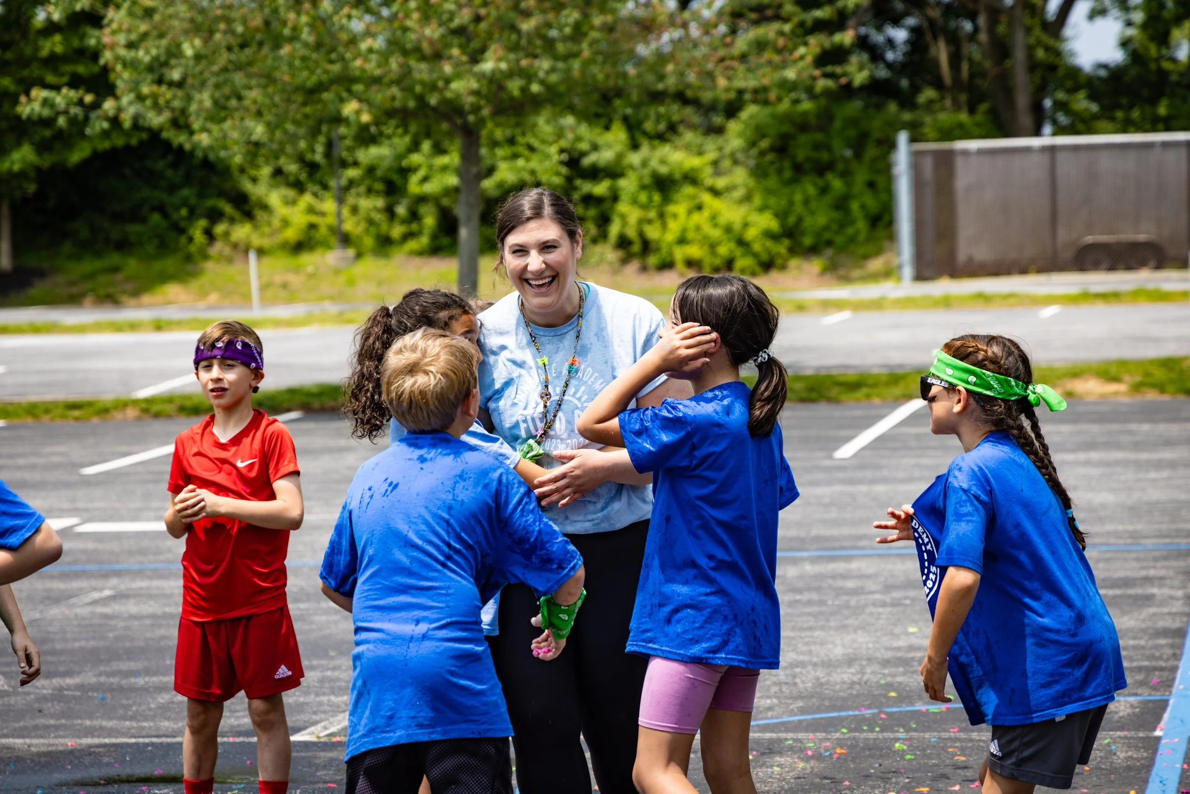 A group of children and a woman laughing and talking outdoors on a sports field, with children wearing colorful bandanas and sports uniforms.
