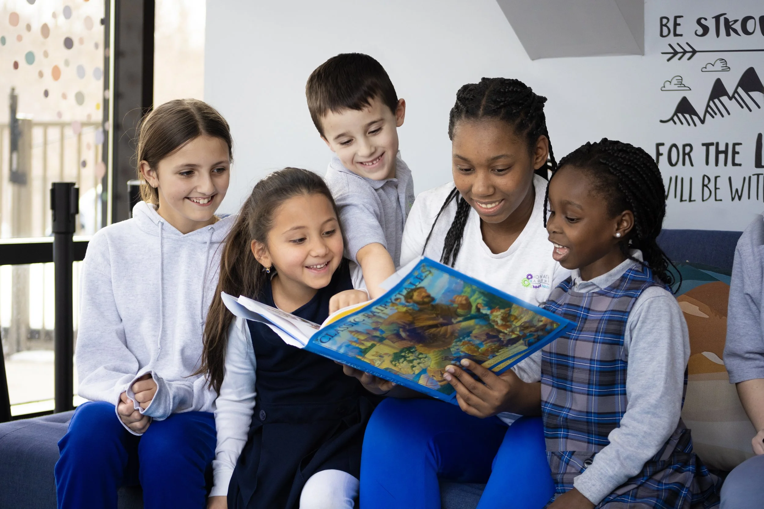 A group of children and a woman are sitting together, looking at a colorful book and smiling. The scene appears to be indoors in a bright classroom or reading area.