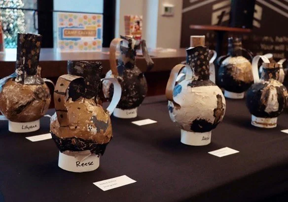 Collection of decorated clay or ceramic vases displayed on a table at an event, each labeled with different names.