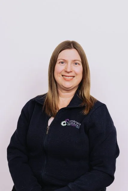 A woman with shoulder-length blonde hair, wearing glasses, a black jacket with a logo that says 'Innovate Academy' and a lanyard with keys, smiling in front of a white background.