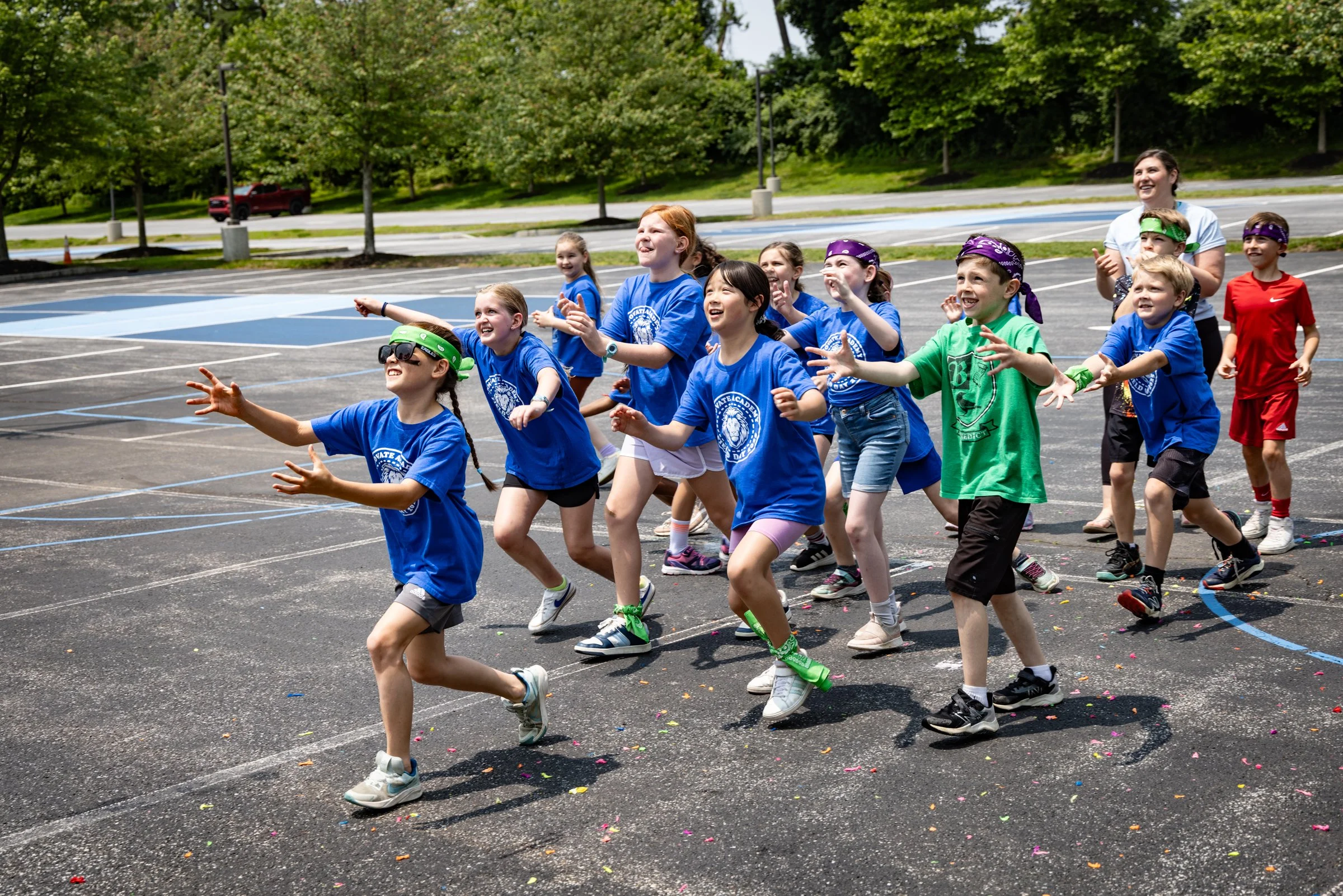Group of children participating in an outdoor event, running and smiling on a parking lot with trees and a parking lot in the background.