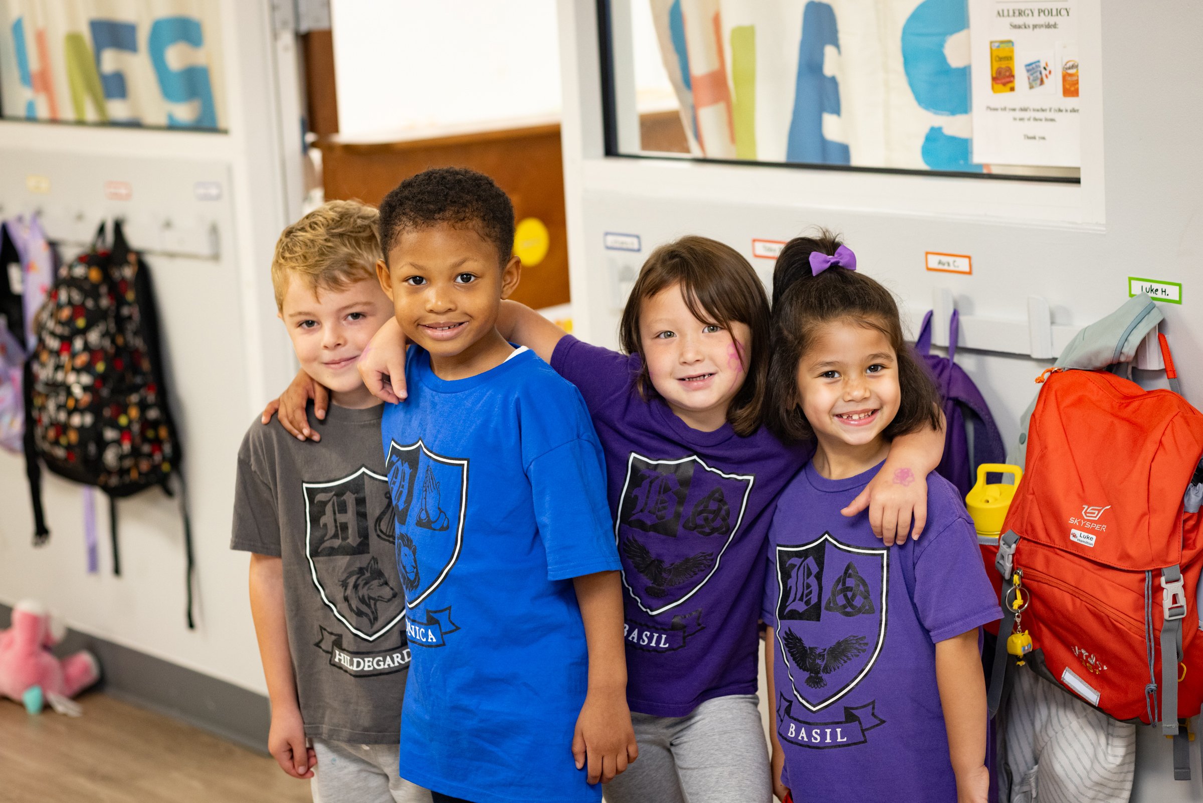 Four young children standing together inside a classroom, smiling and posing for the photo. They are wearing colorful t-shirts with a school emblem, and there are backpacks hanging on hooks behind them.