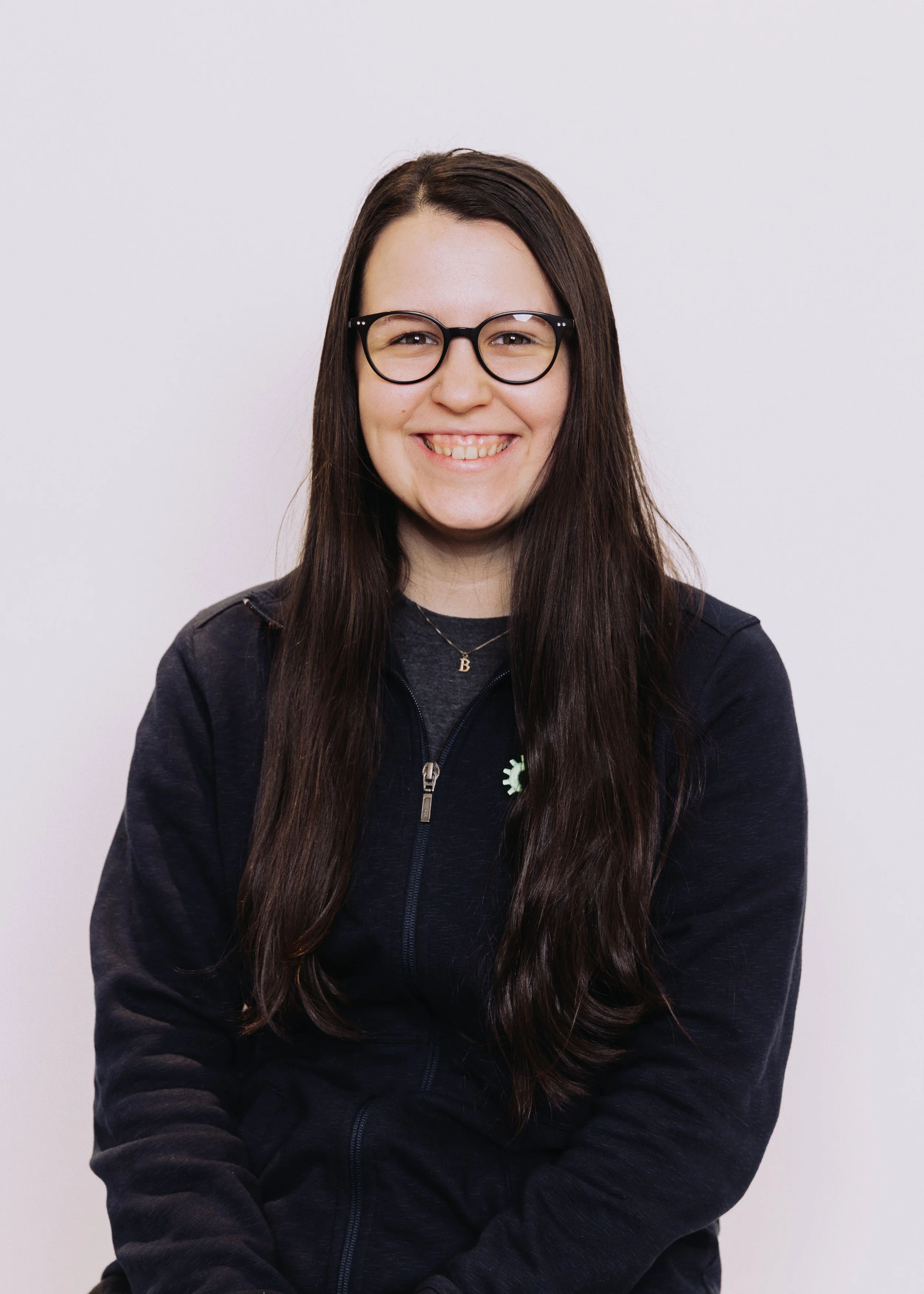 Portrait of a smiling woman with long dark hair, glasses, and casual dark clothing standing against a plain white background.