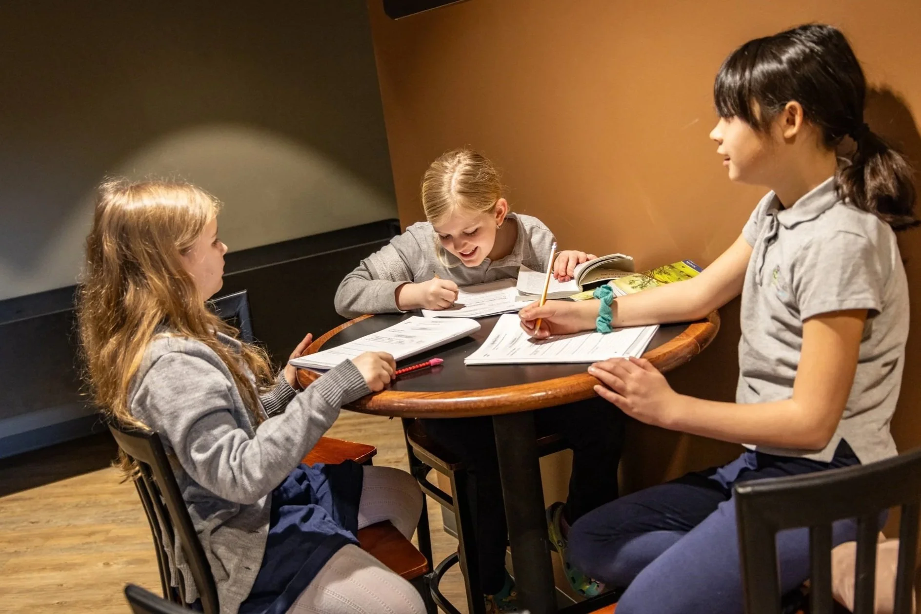 Four young girls sitting around a table, engaged in a lively conversation or activity, with books and notebooks open in front of them.