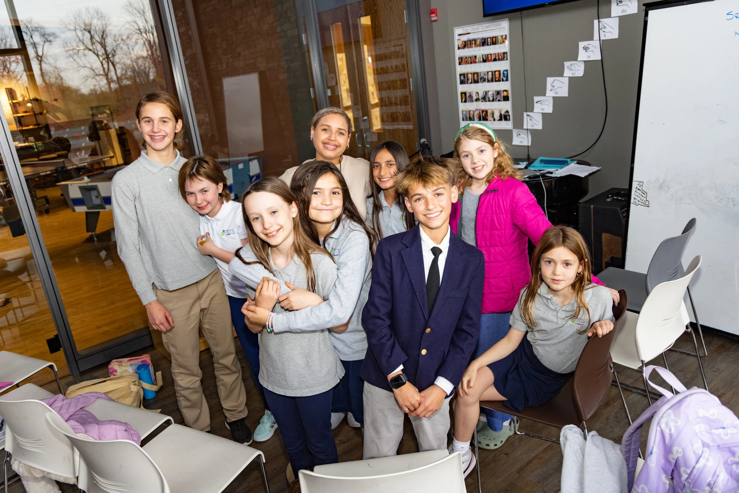 A group of smiling children and a teacher posing in a classroom with chairs, backpacks, and classroom decorations in the background.