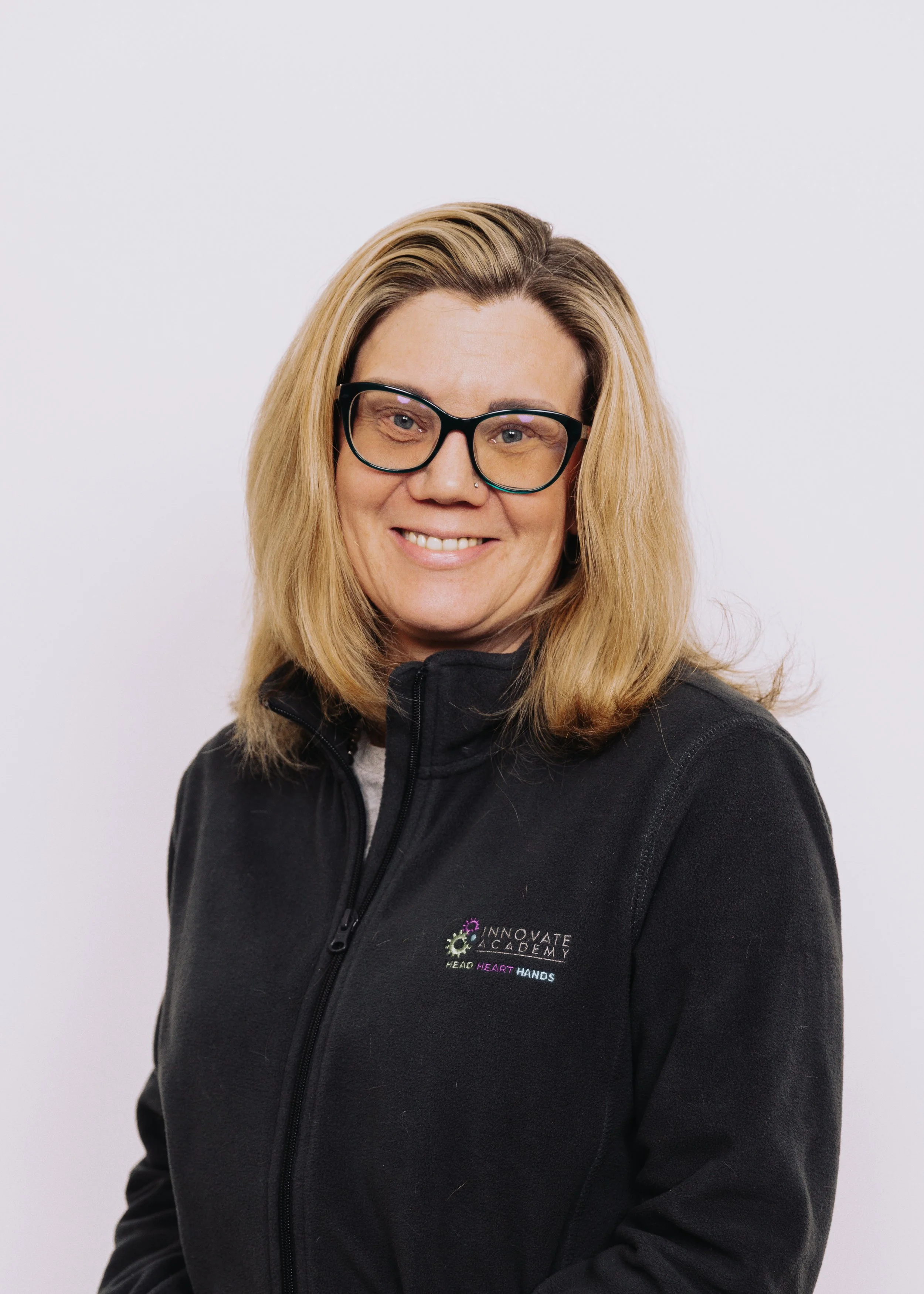 Woman with blonde hair, glasses, and a black jacket with a logo that says 'Innovate Academy' and 'Head, Heart, Hands,' smiling against a plain white background.