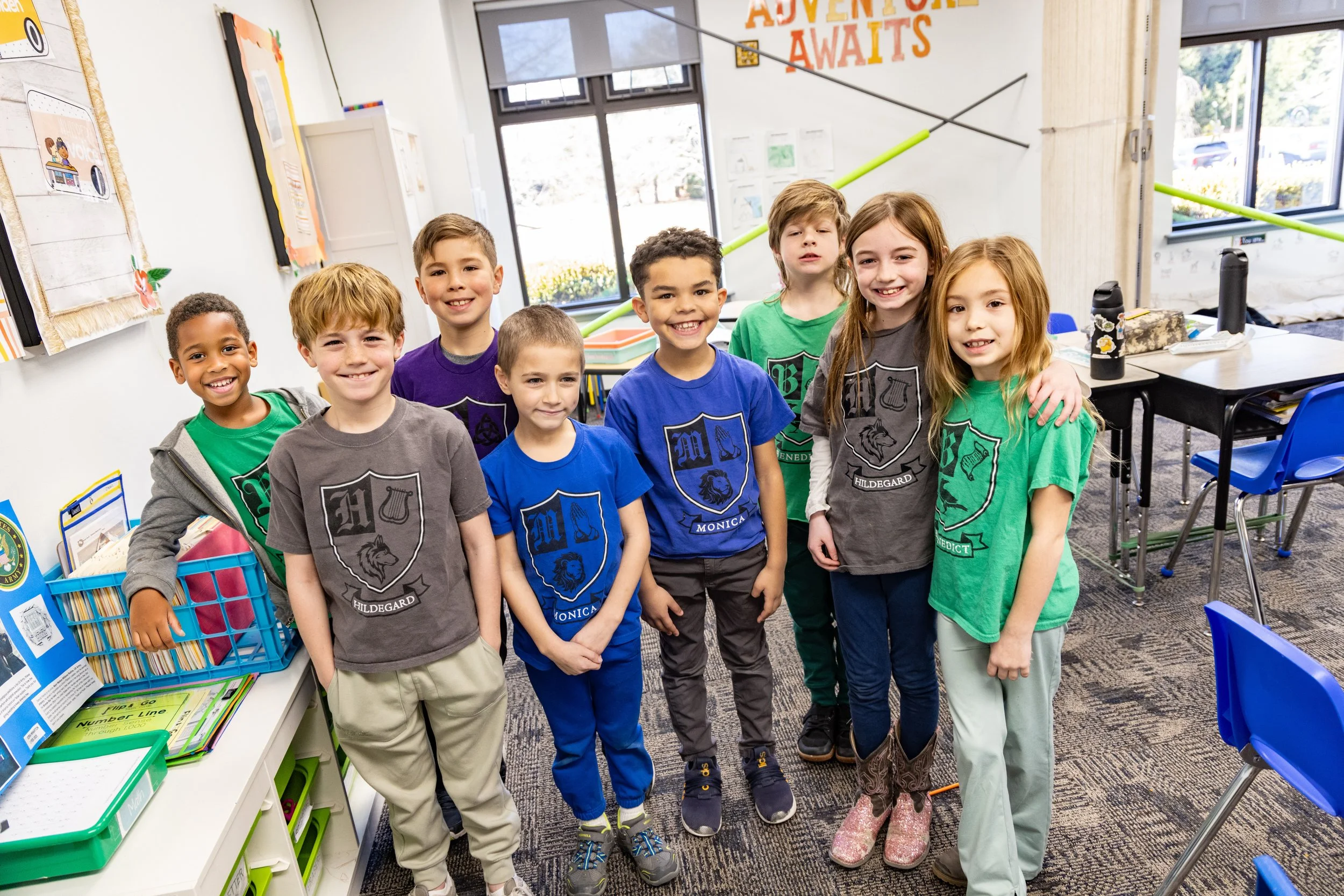 Group of eight children smiling and standing together in a classroom, wearing colorful t-shirts with school logos, near school supplies and desks.