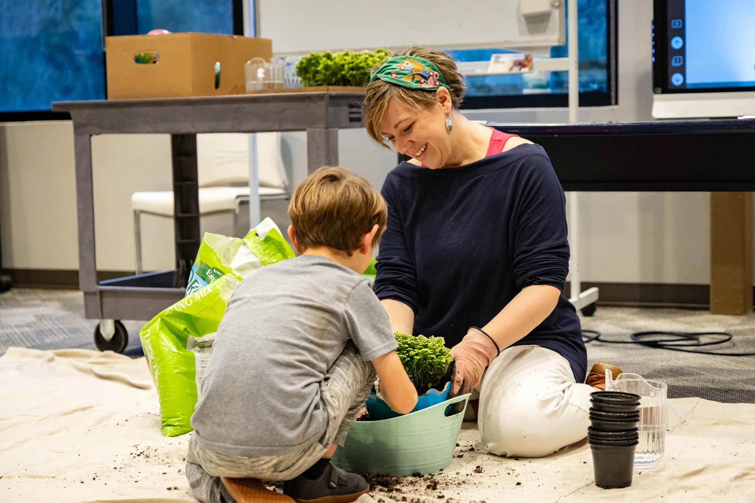 A woman and a young boy gardening together indoors, sitting on a white sheet with gardening supplies around them, smiling as they plant green plants.