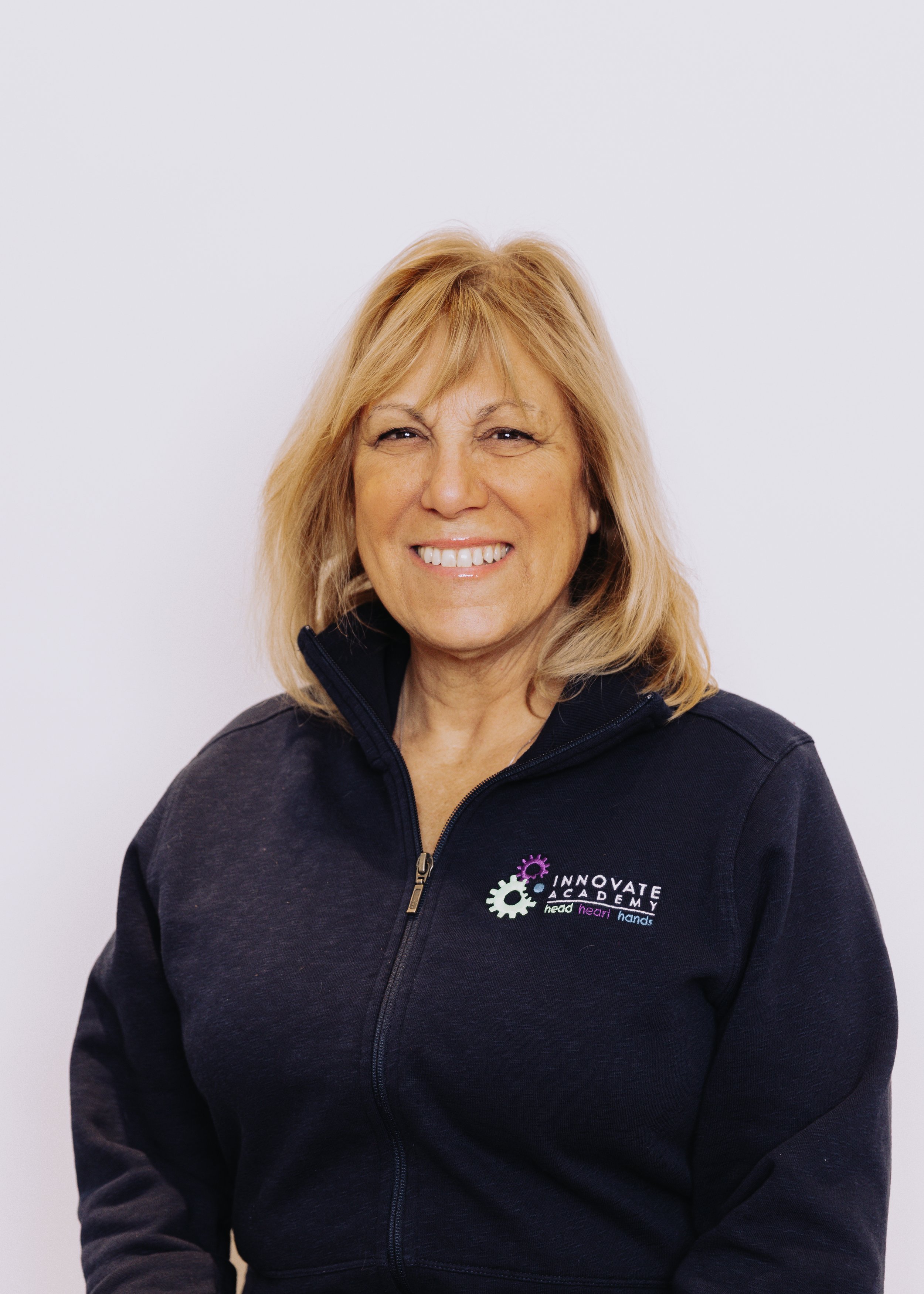 Smiling woman with blonde hair wearing a black zip-up jacket with 'Innovate Academy' logo on chest, standing against a plain white background.