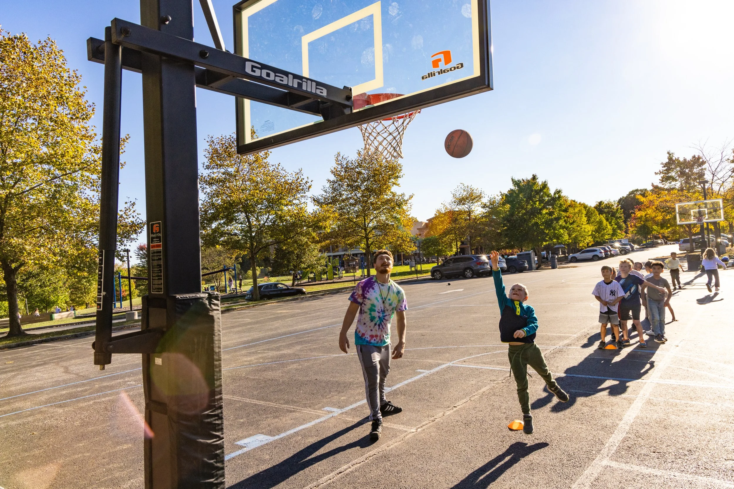 Children playing basketball outdoors on a sunny day at a park gymnasium.