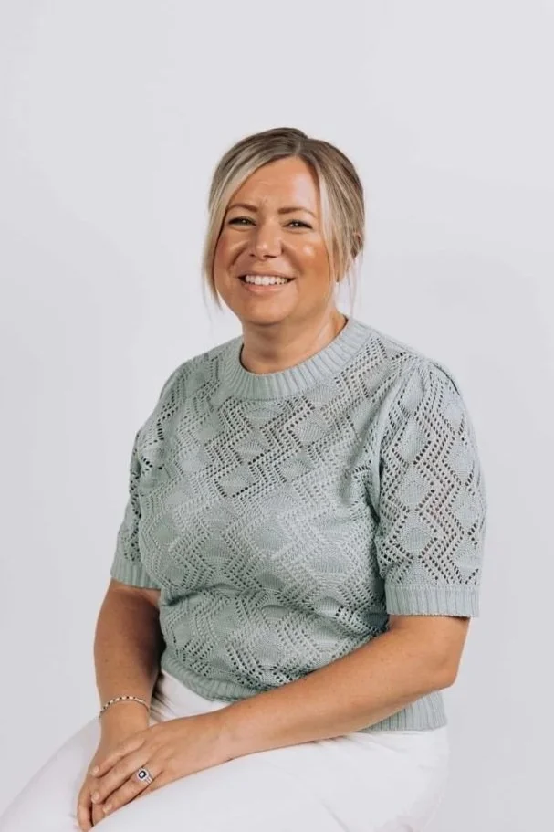 A woman with blonde hair smiling while sitting, wearing a light blue lace-patterned sweater and white pants, against a plain white background.
