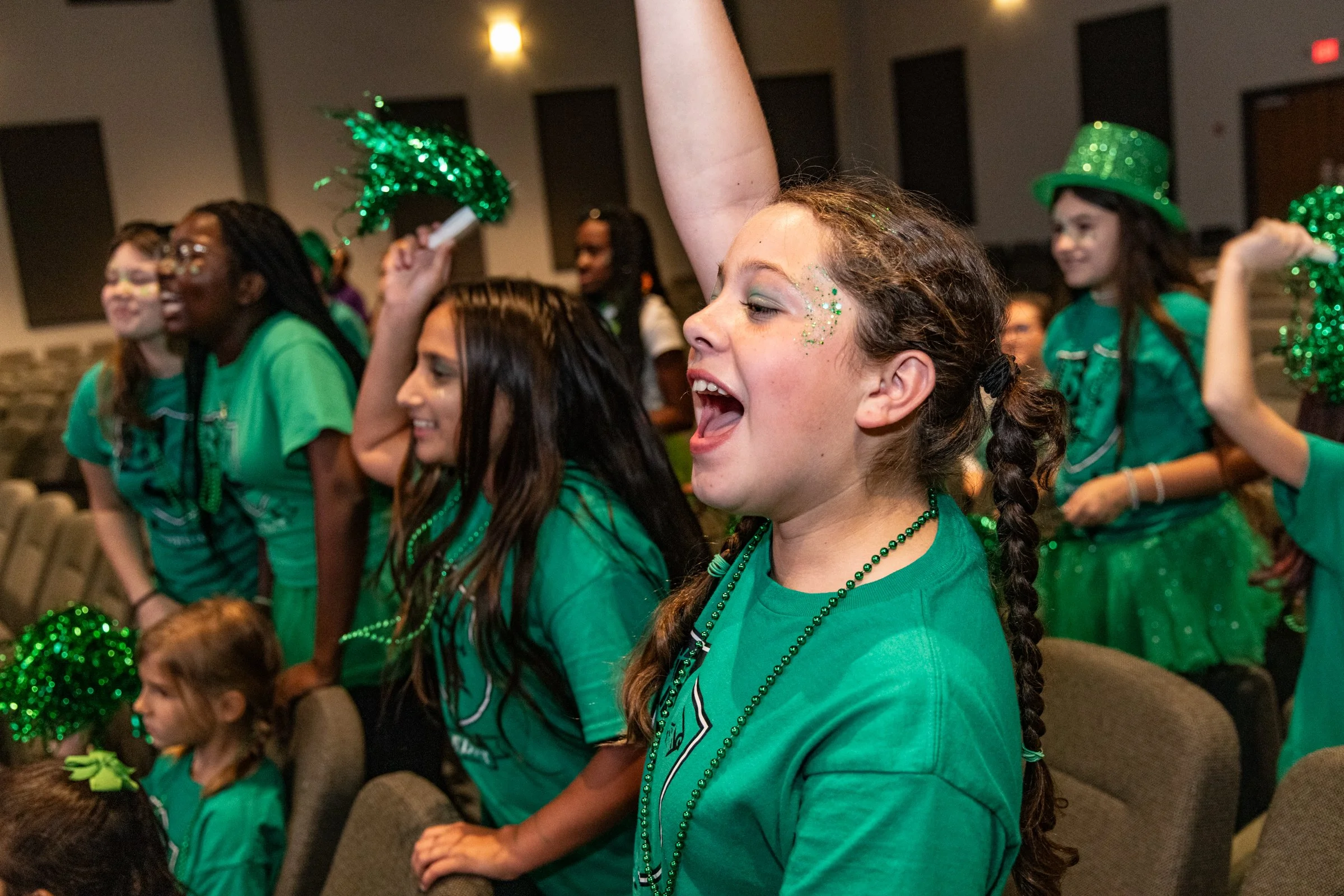 Group of young girls celebrating, wearing green shirts, with some holding green pom-poms and wearing green glittery top hats, in a festive indoor setting.
