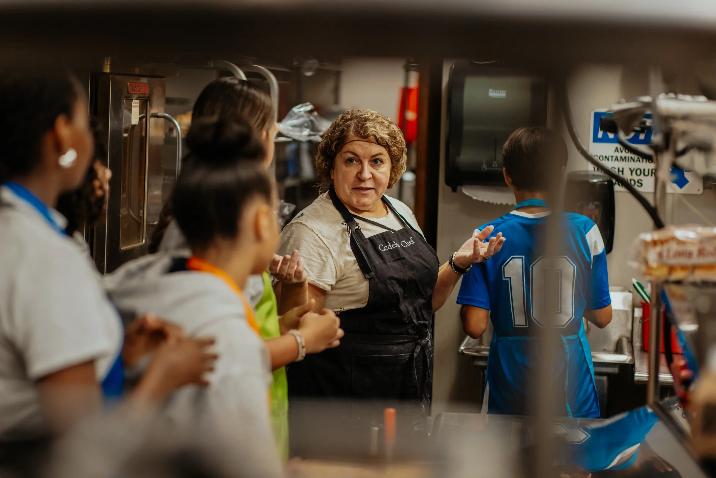 A female chef speaking to a group of children in a kitchen setting.