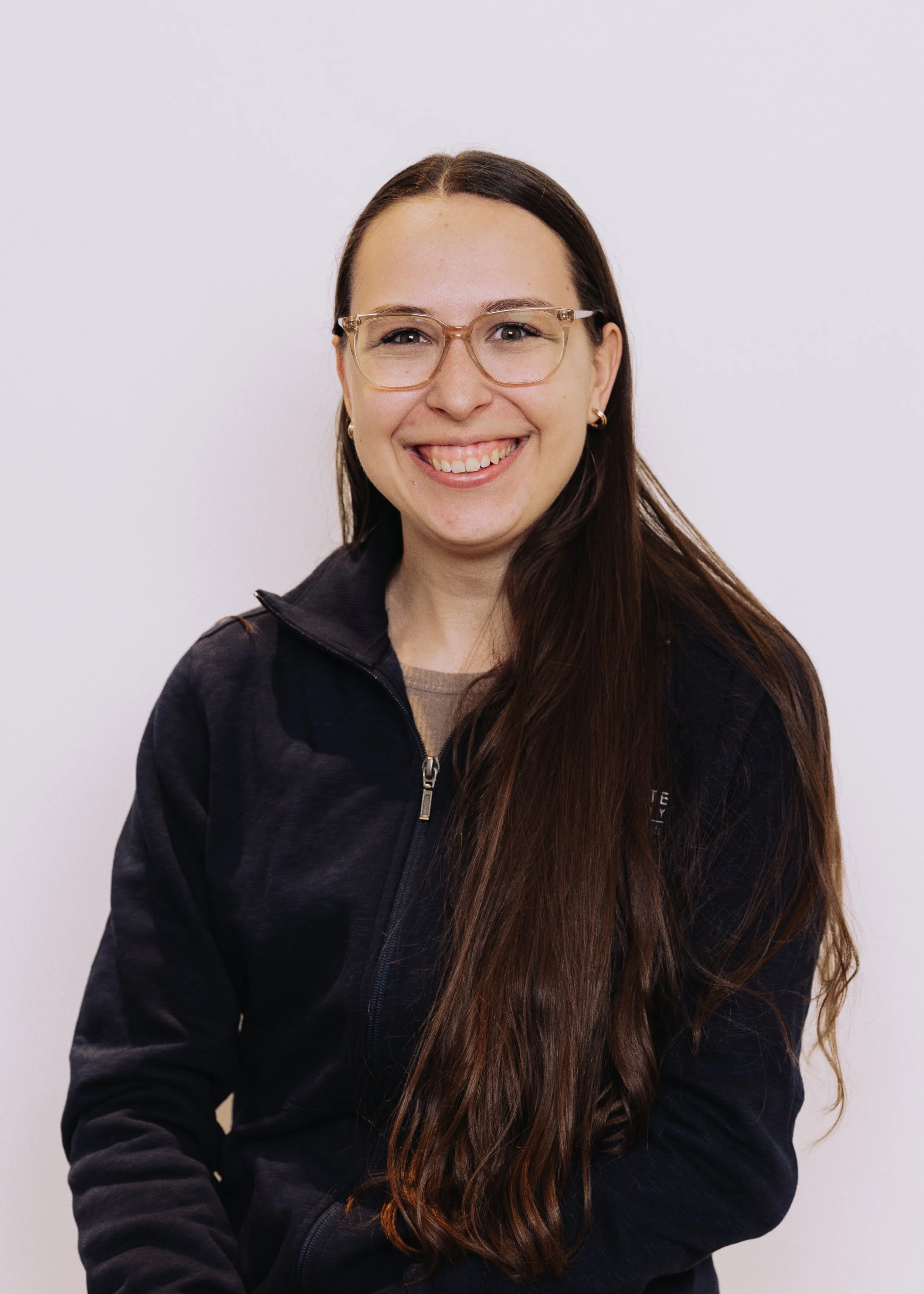 Smiling woman with long brown hair, glasses, and gold earrings, wearing a dark zip-up jacket, against a plain white background.