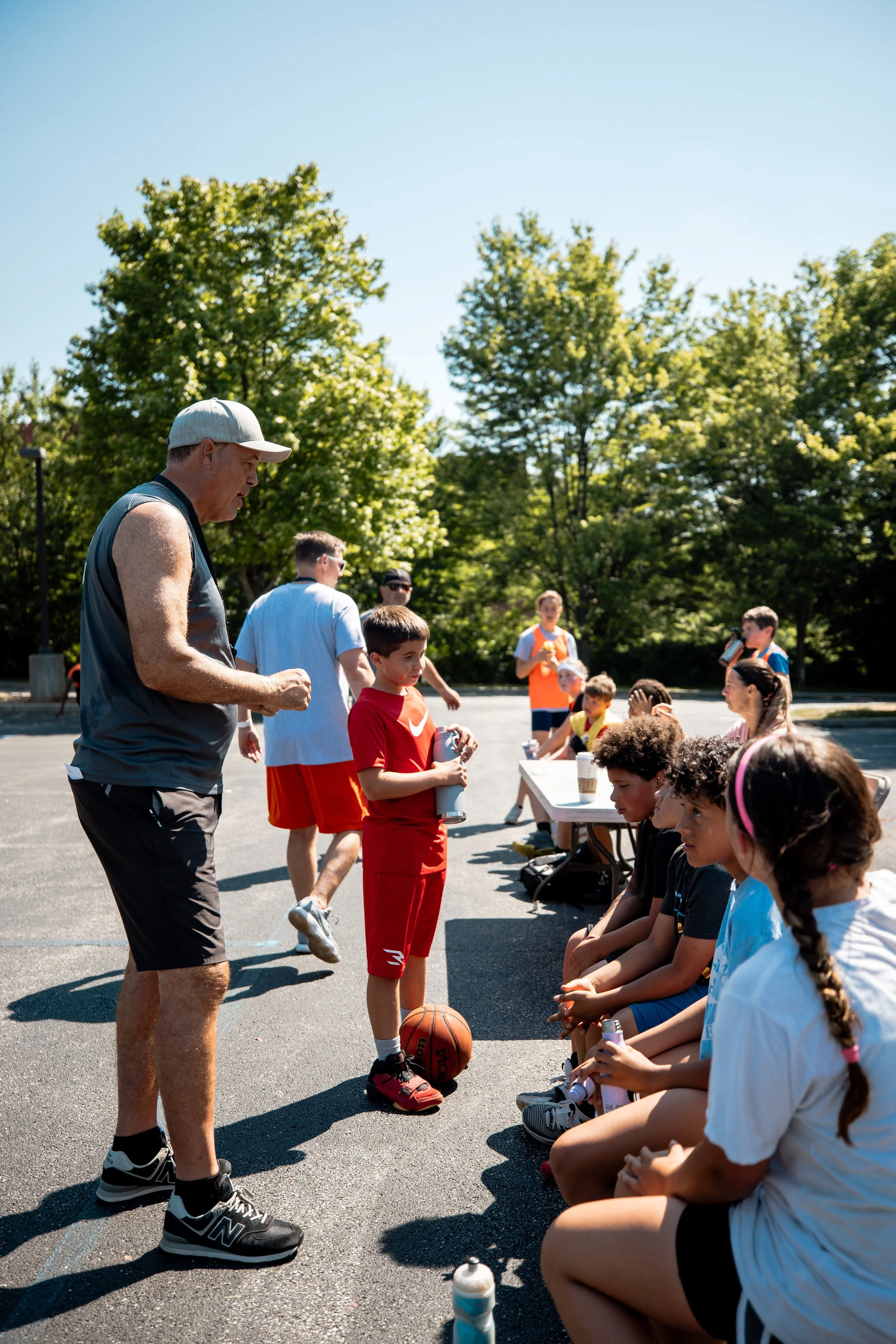 A youth basketball coach addresses a group of children sitting on a bench during a sunny outdoor practice or game, with trees and a clear sky in the background.