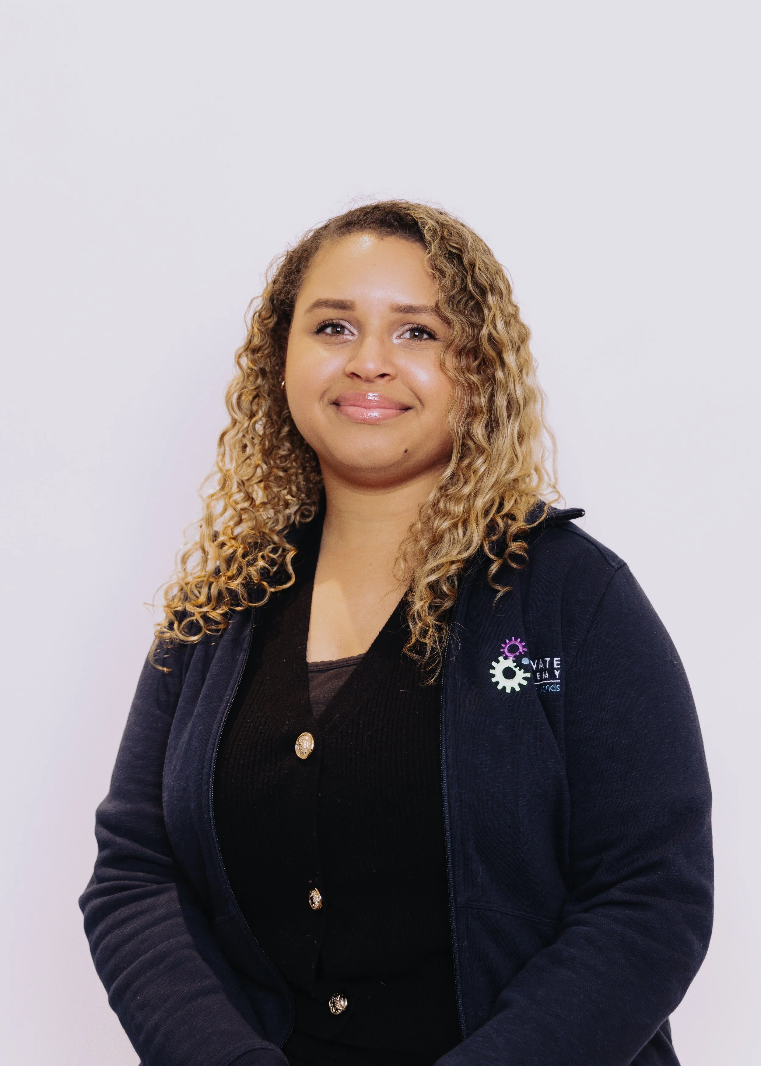 A woman with curly blonde hair, wearing a black top and a navy zip-up jacket with a logo on the sleeve, standing against a plain white background and smiling gently.