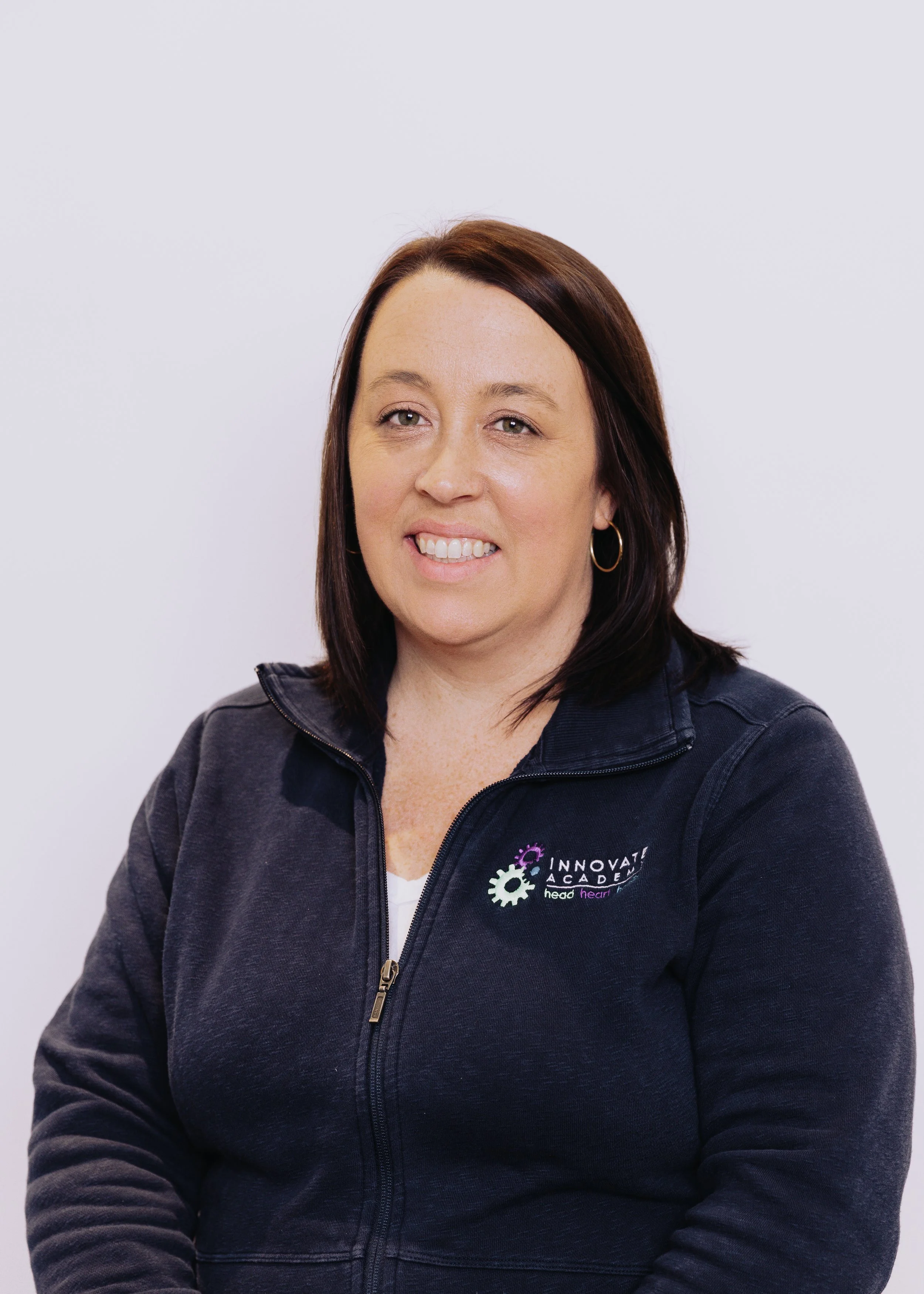 A woman with shoulder-length brown hair and hoop earrings, wearing a navy zip-up jacket with an 'Innovate Academy' logo on the chest, standing against a plain white background.