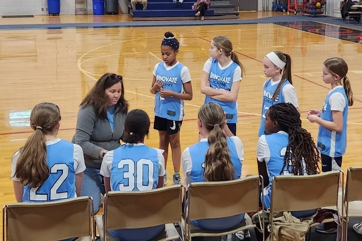 A youth basketball team having a timeout on an indoor court. The coach is talking to the players who are sitting and standing, wearing blue and white uniforms with numbers.