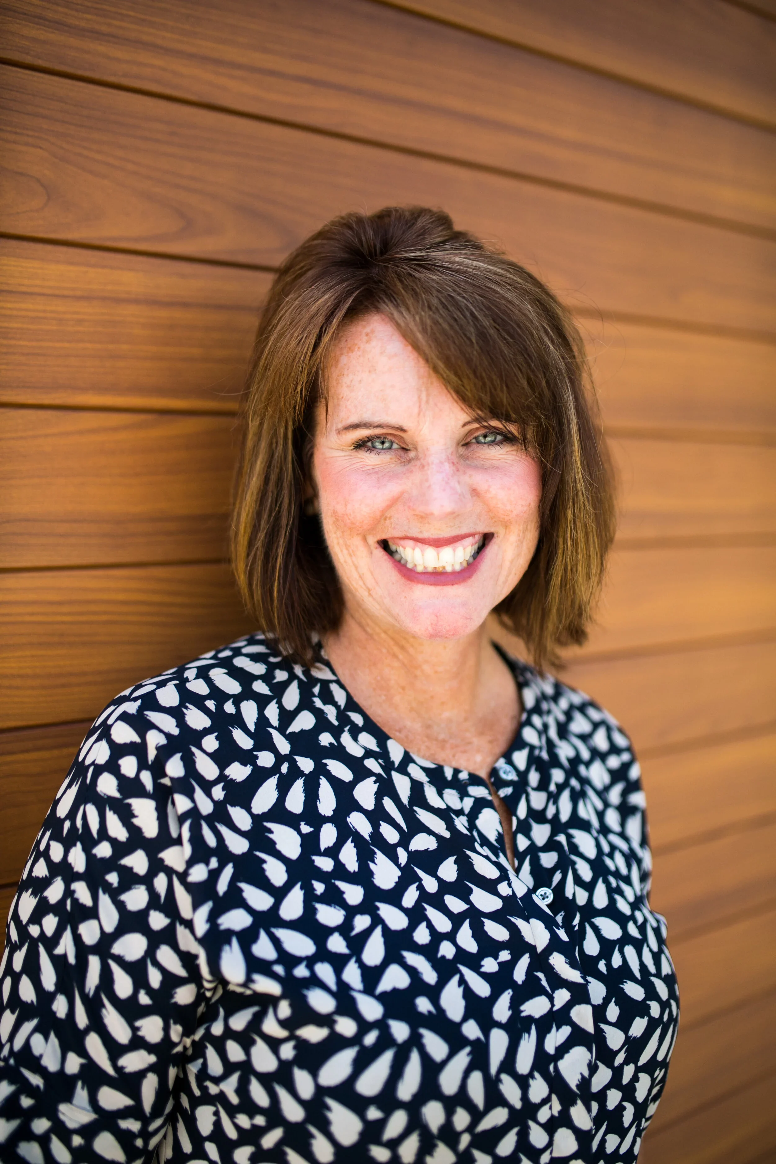 A smiling woman with shoulder-length brown hair and blue eyes, wearing a black and white patterned blouse, standing against a wooden wall.