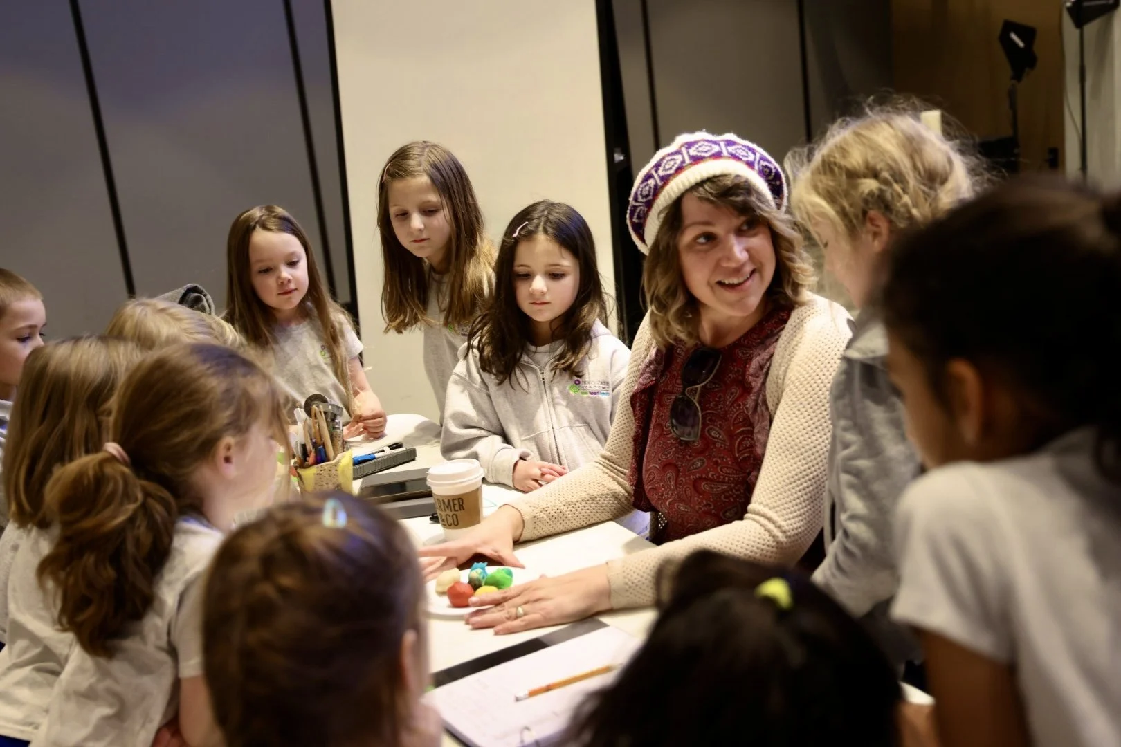 A woman wearing a colorful knitted hat is sitting at a table with young girls, showing them small brightly colored objects. The girls are gathered around, looking at what she is showing.
