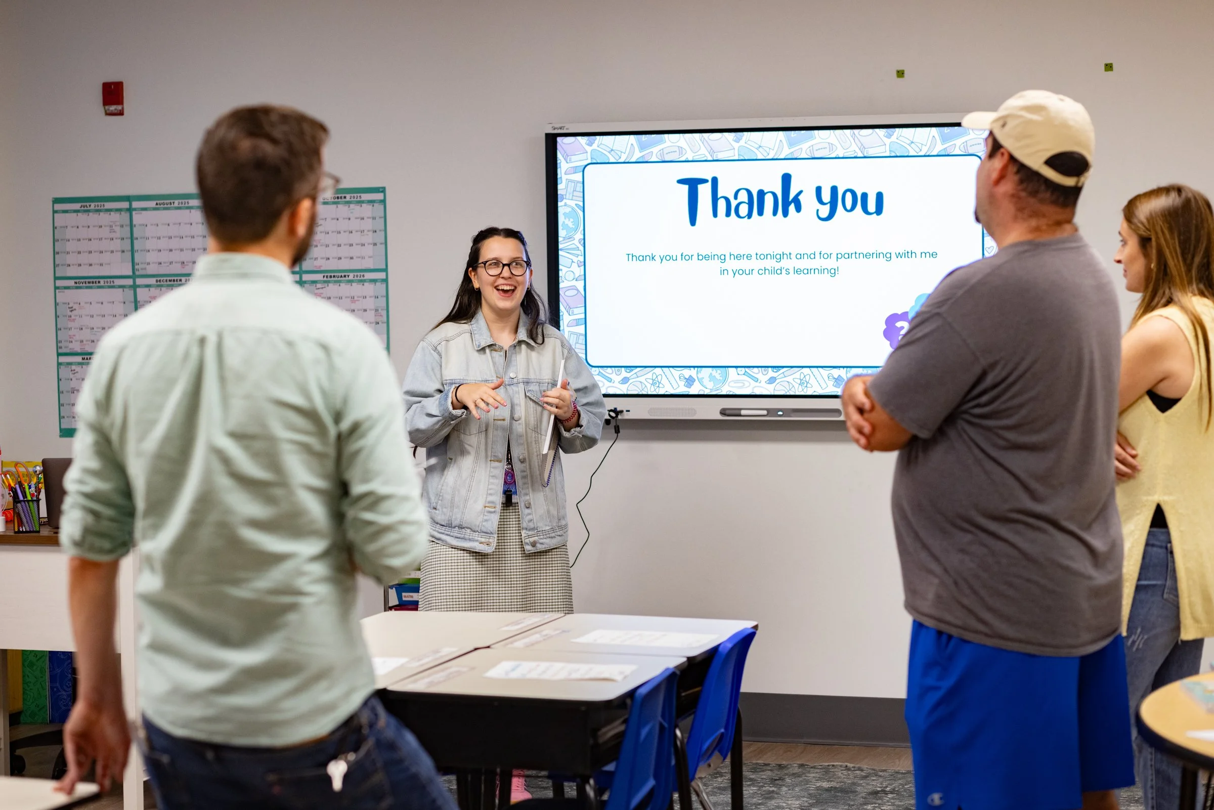 Teacher smiling and speaking to a group of four adults in a classroom, with a large screen displaying a thank you message