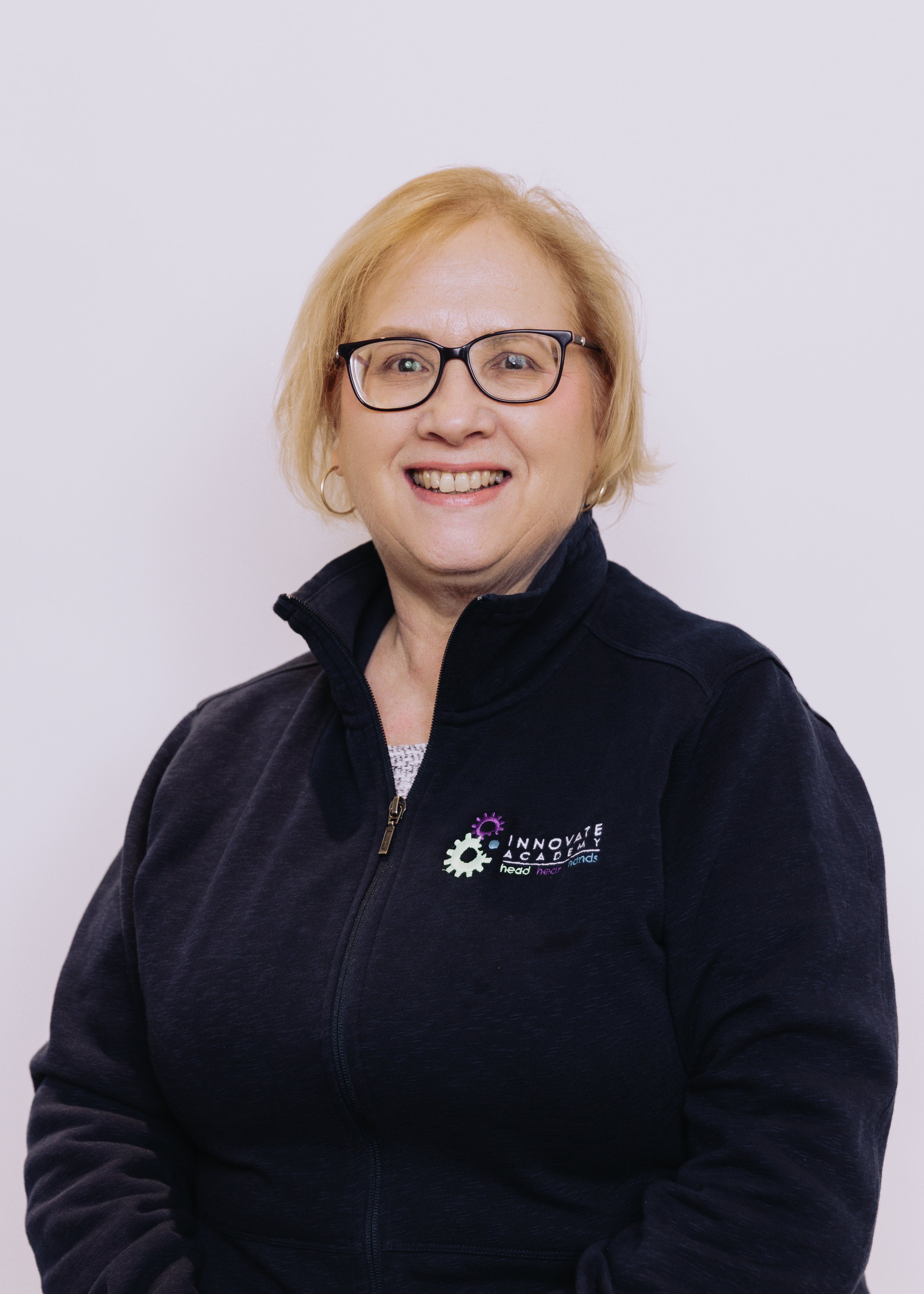 A woman with blonde hair, glasses, and a black jacket with a logo that reads "INNOVATE ACADEMY" and a phrase "head neck hands". She is smiling against a plain white background.