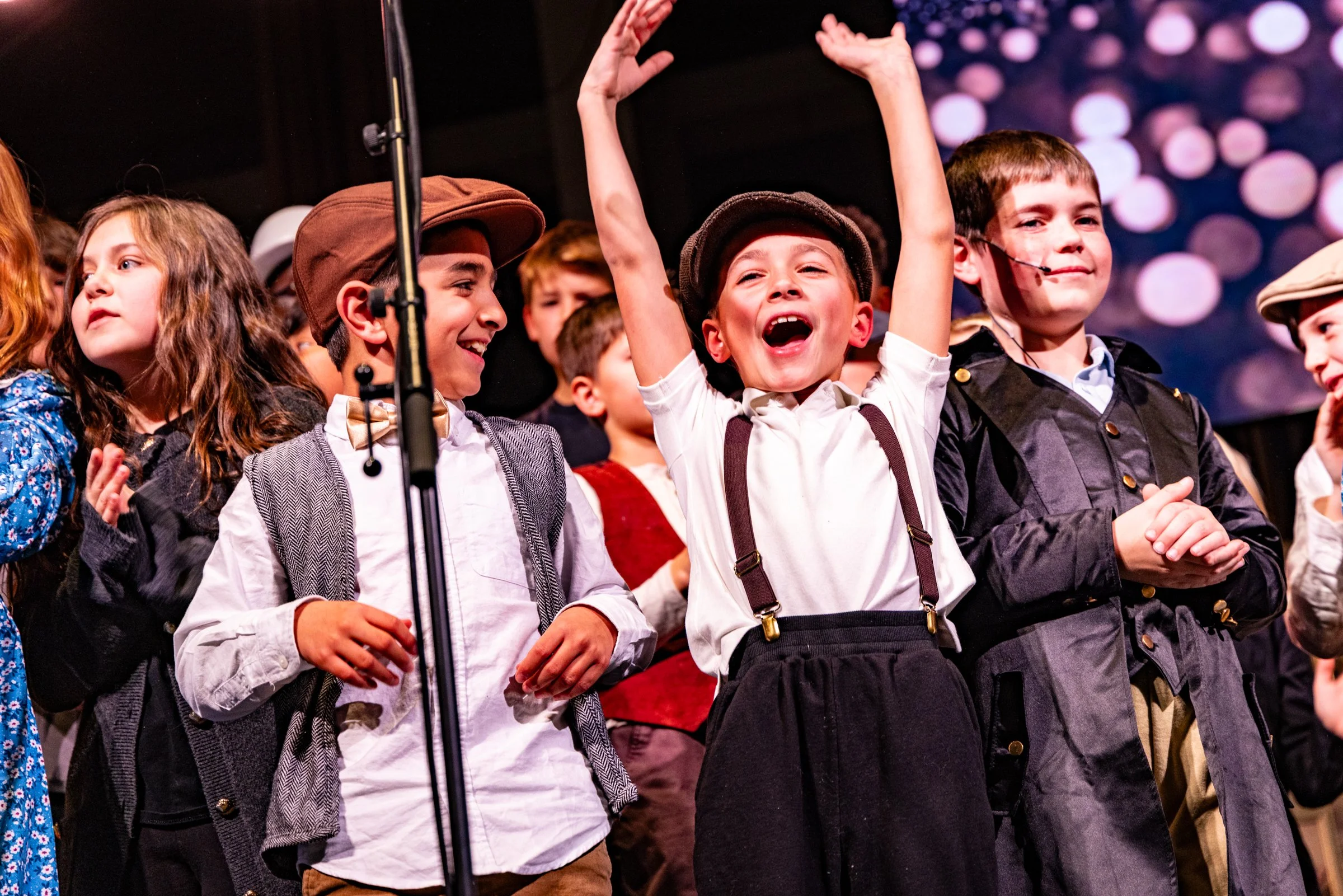 Children dressed in vintage clothing singing or performing on stage with a microphone, smiling and cheering.