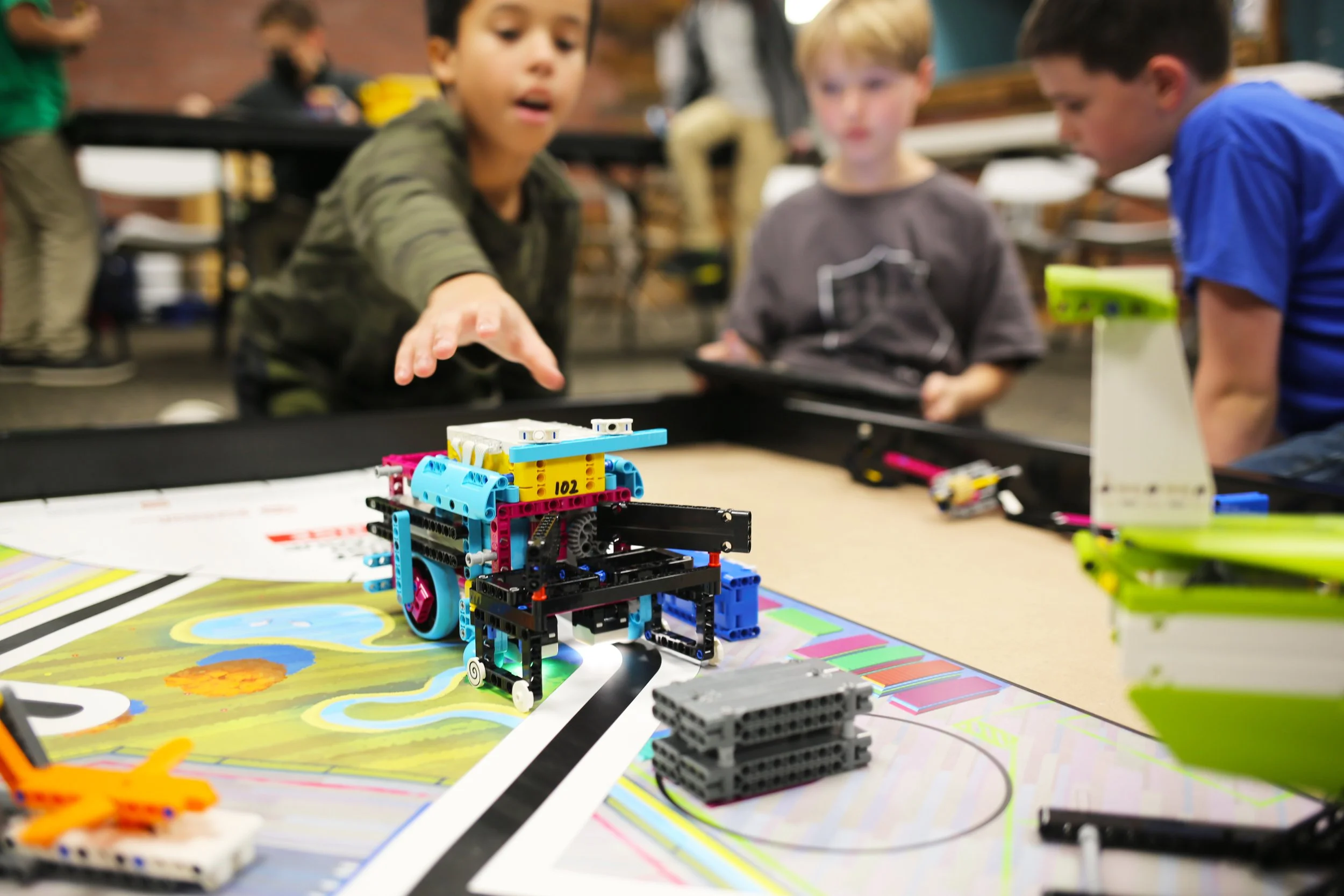 Group of children gathered around a table, observing a colorful LEGO robot on a game board, with some children using tablets.
