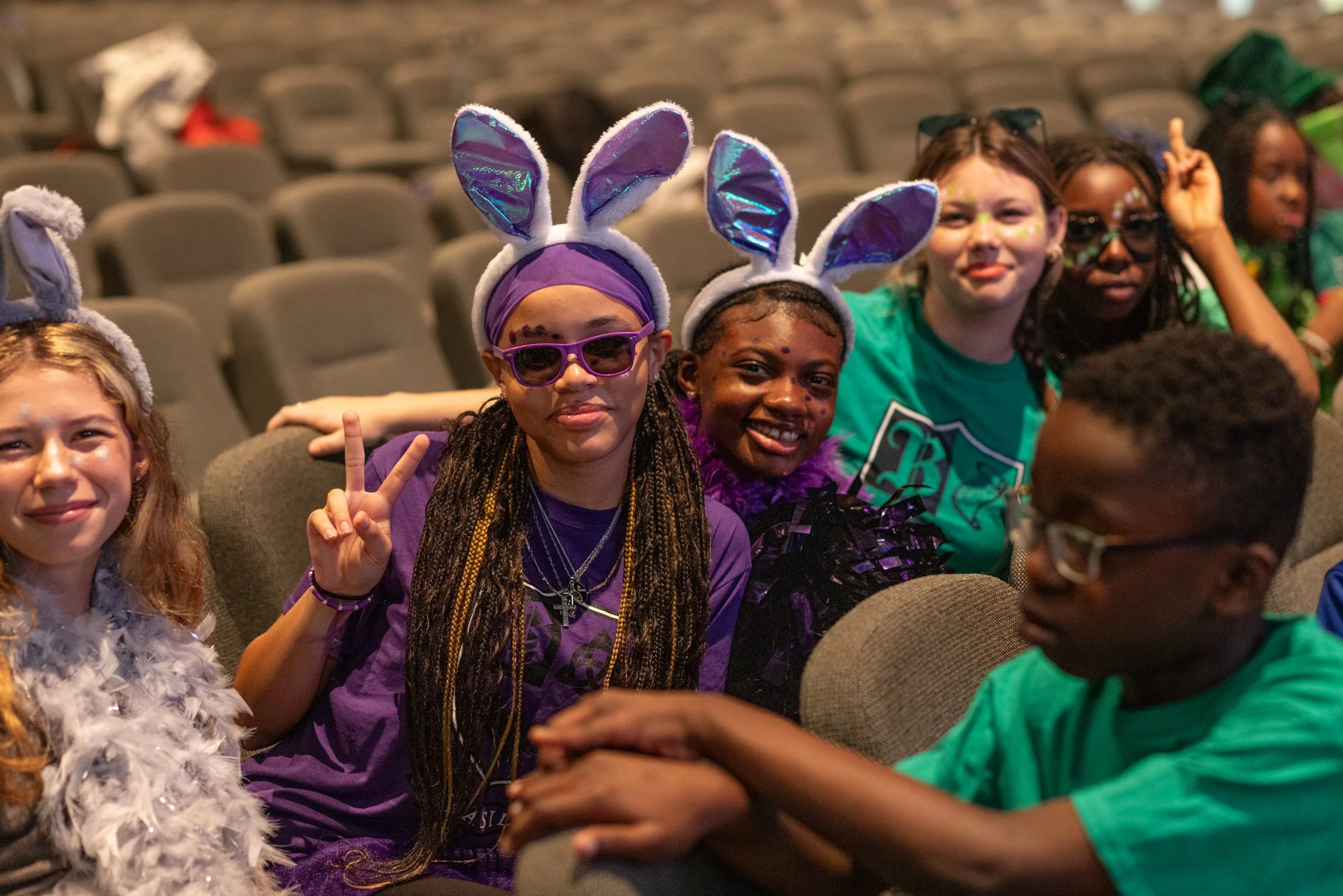 Group of children dressed in colorful costumes, wearing bunny ears, sitting in an auditorium.