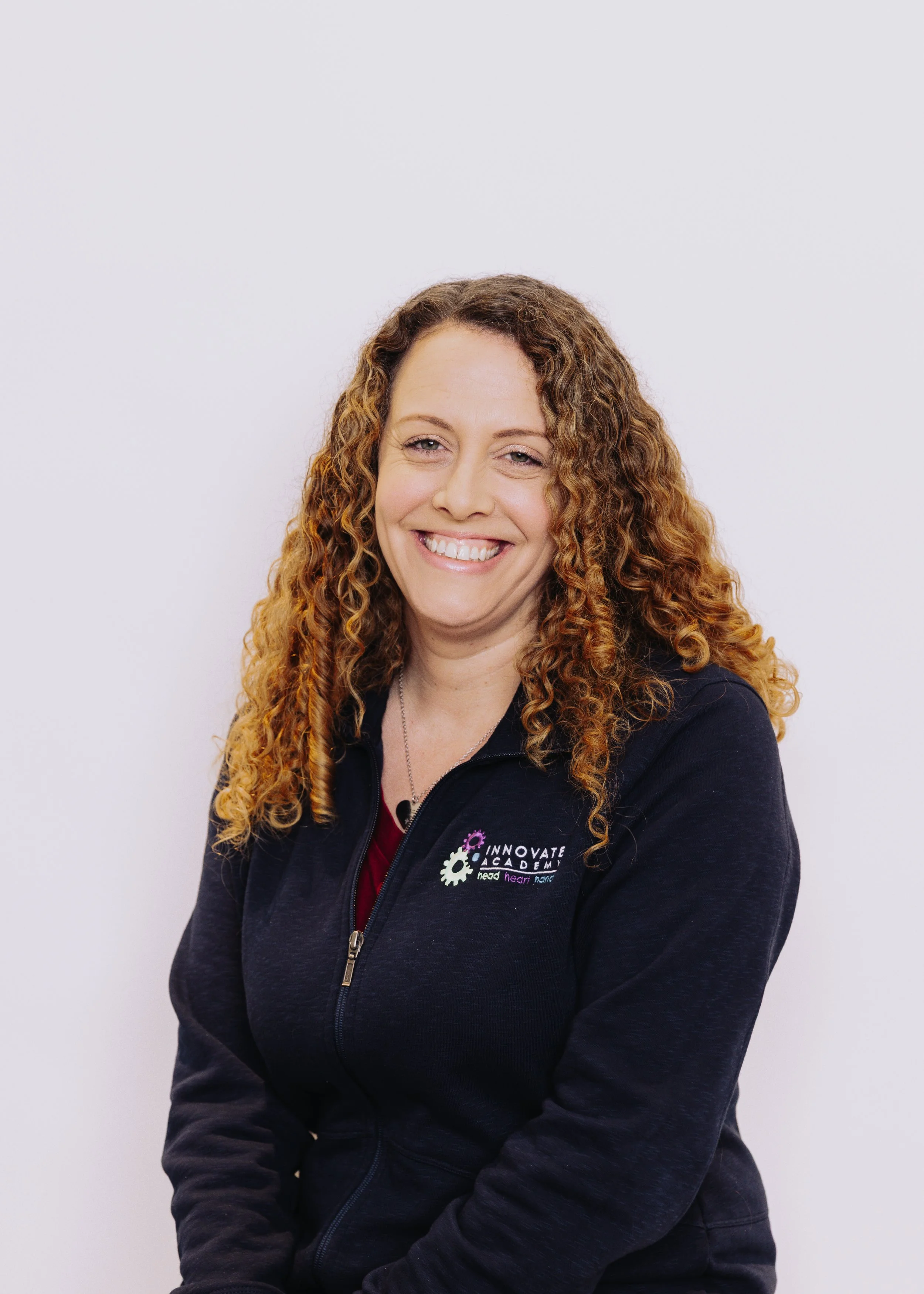 A woman with curly red hair smiling at the camera, wearing a dark zip-up jacket with a logo that says 'Innovate Academy' and the words 'head, hear, move' underneath.