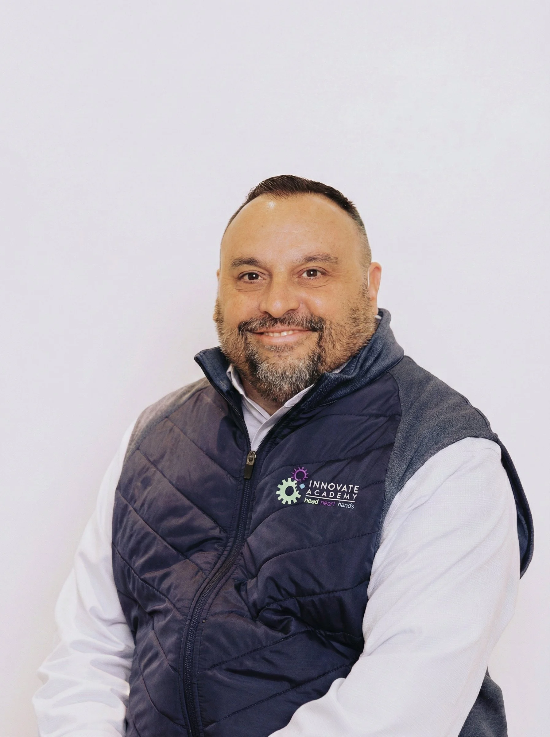 A man smiling, wearing a white shirt and a dark vest with the logo 'Innovate Academy' on it, sitting against a plain white background.