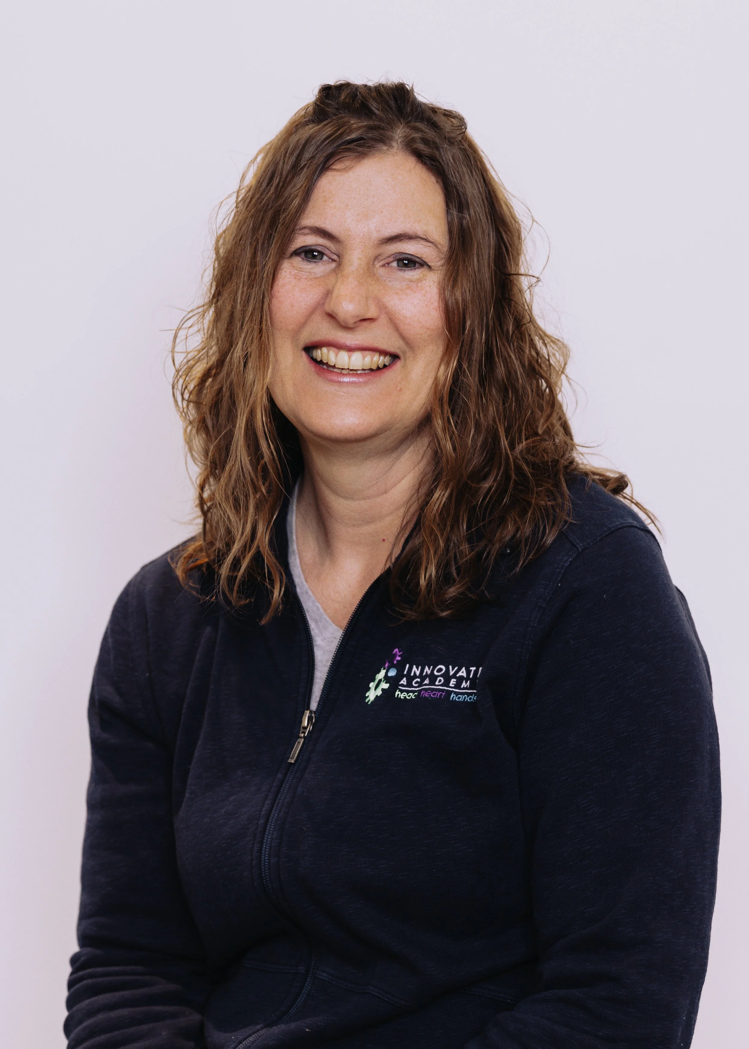 A smiling woman with curly brown hair wearing a navy blue zip-up jacket with the logo 'Innovation Academy' on the chest, seated against a plain light background.