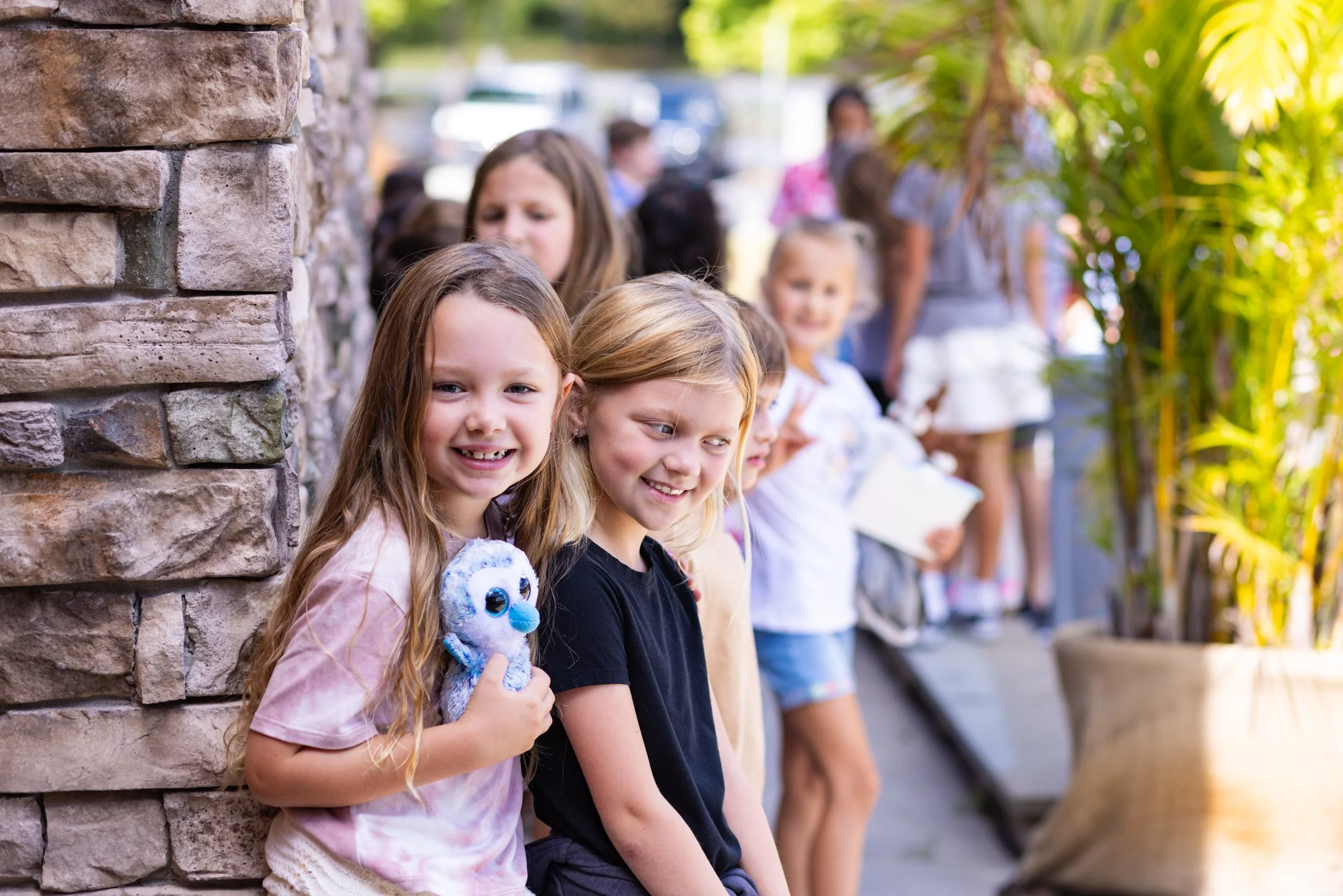 Smiling young girls standing in a line outdoors, waiting, with some girls holding notebooks and a plush toy.