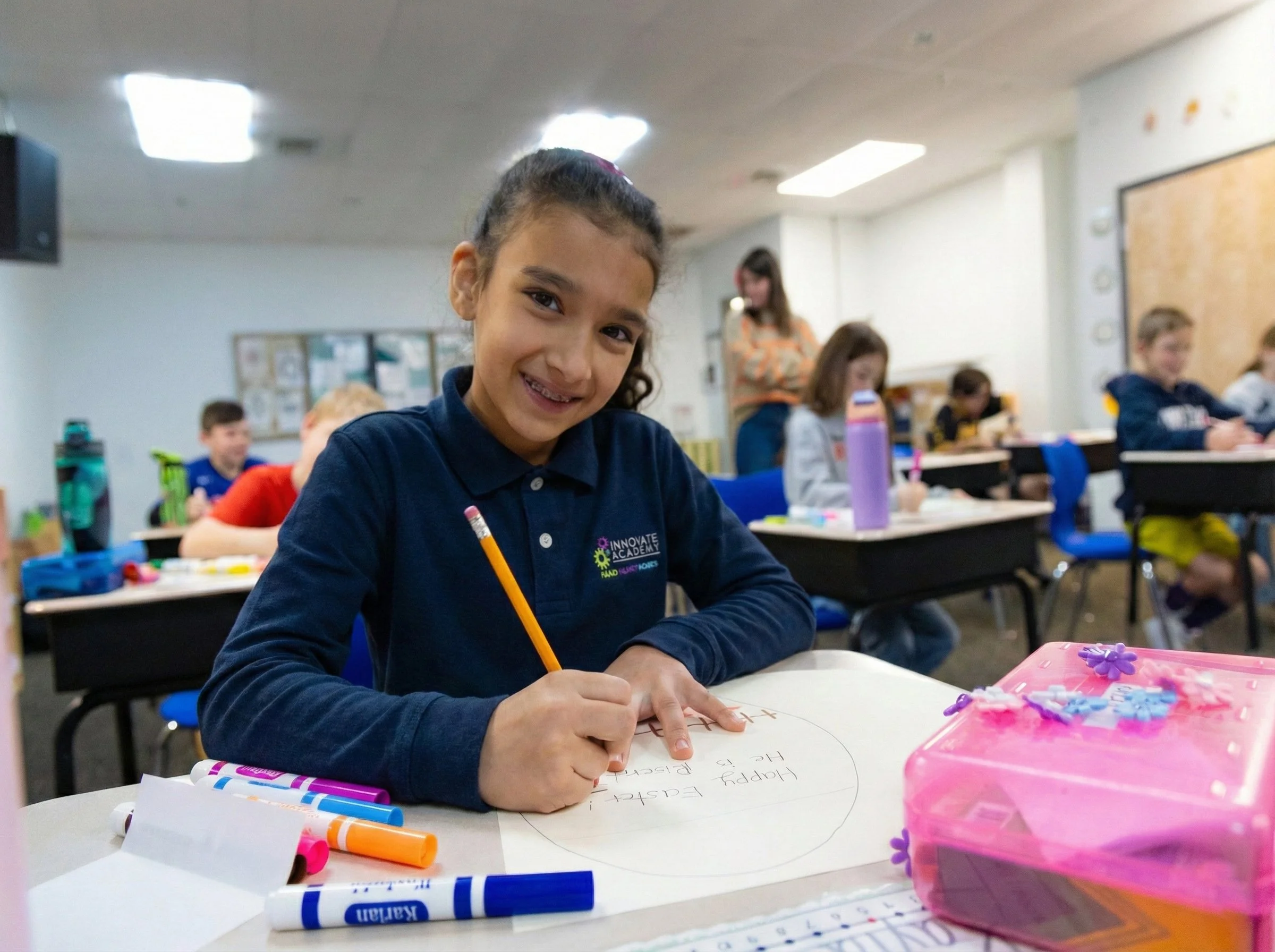 A young girl in a navy blue shirt sits at a desk, smiling while writing on a piece of paper, in a classroom filled with other children and a teacher in the background.